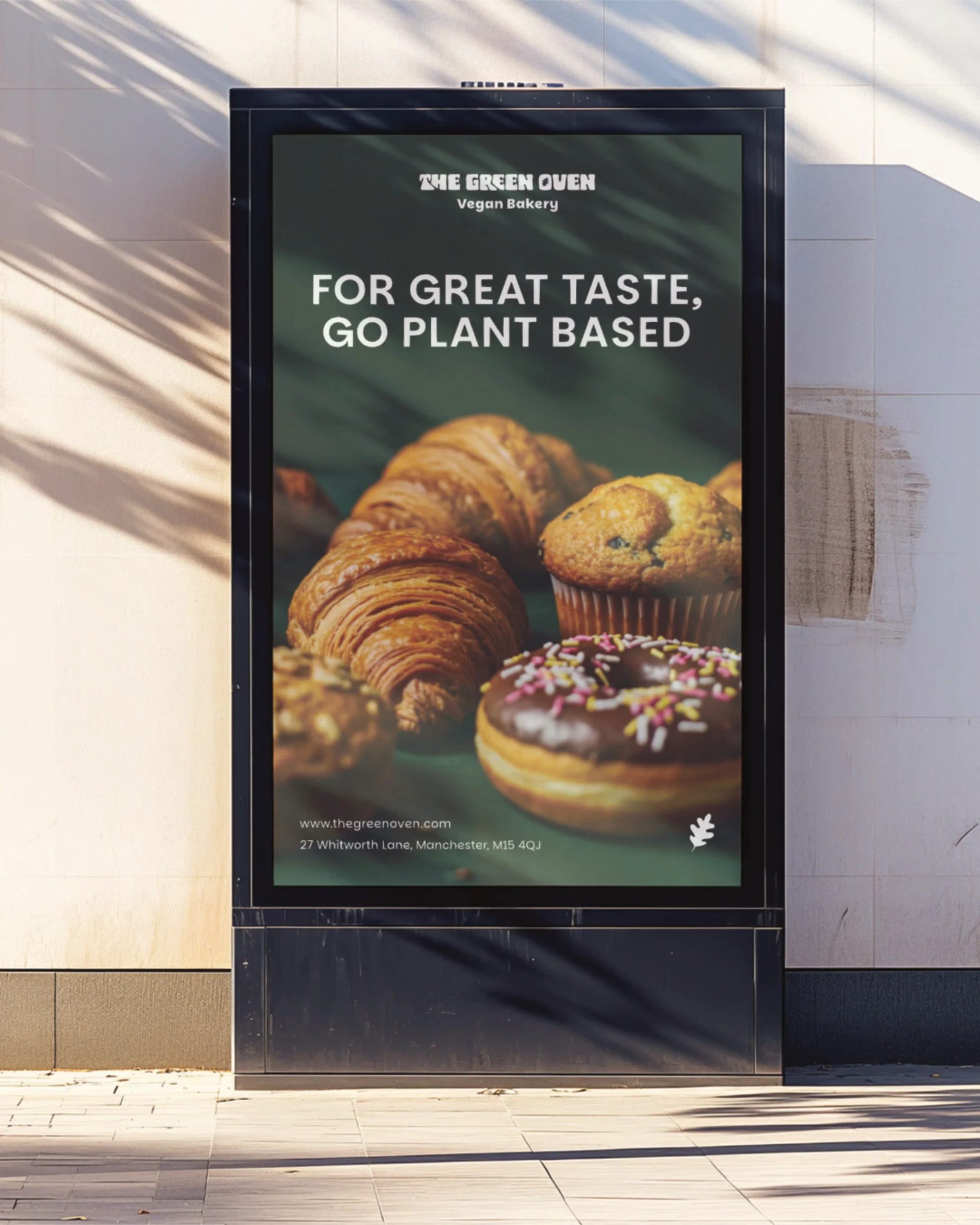 An outdoor advertisement for The Green Oven vegan bakery features images of croissants, muffins, and donuts and includes the text 'For great taste, go plant based.' The ad also provides the bakery's website and address in Manchester.