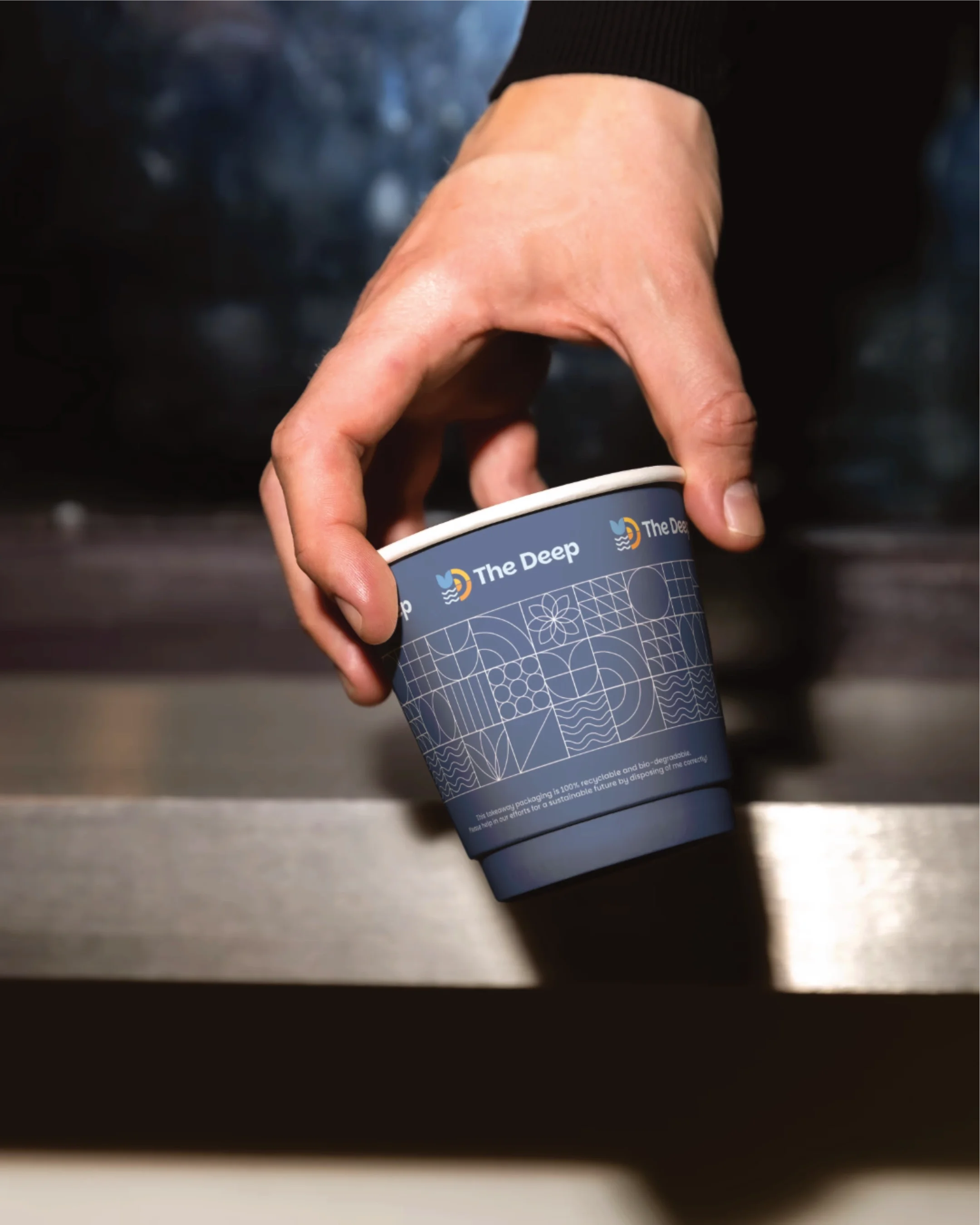 A person's hand holding a blue paper cup with a geometric pattern and the text 'The Deep' on a dark surface.
