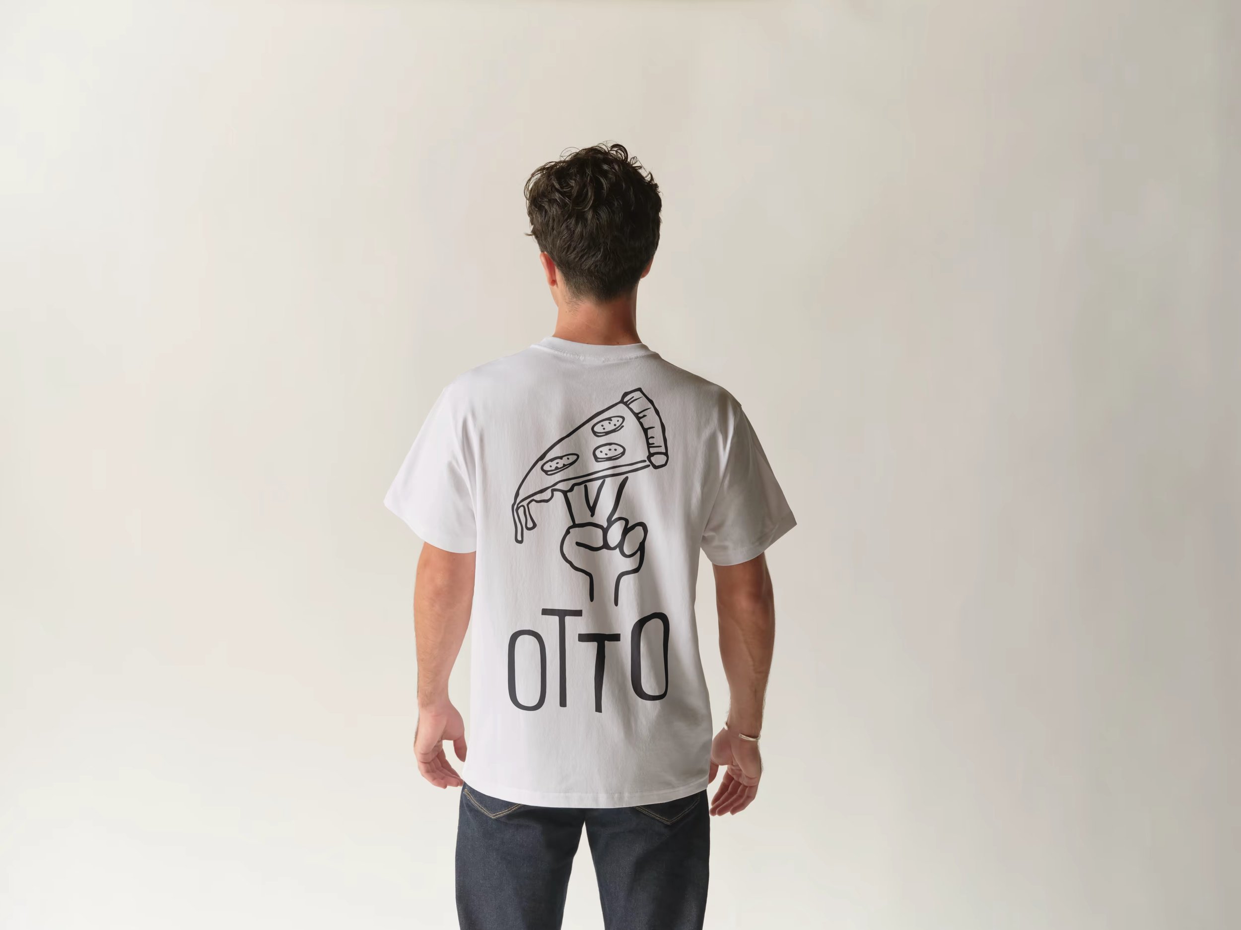 Back view of a person with short dark curly hair, wearing a white T-shirt with a pizza slice graphic and text, standing against a plain off-white wall.