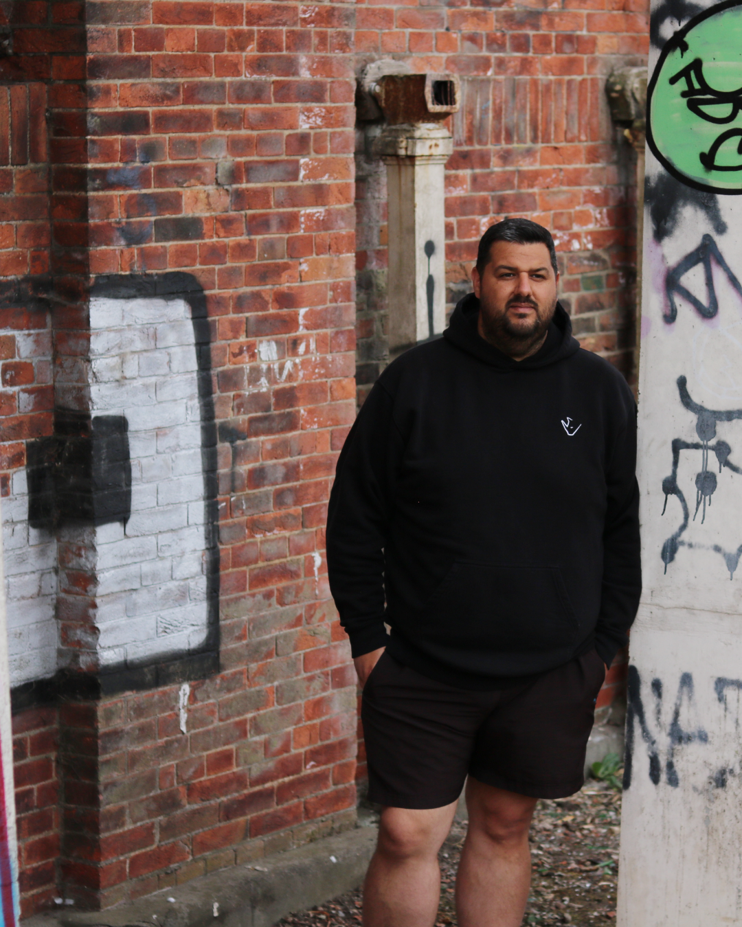 Michael Bullamore standing outdoors next to a graffiti-covered brick wall in Hull wearing a black hoodie and black shorts.