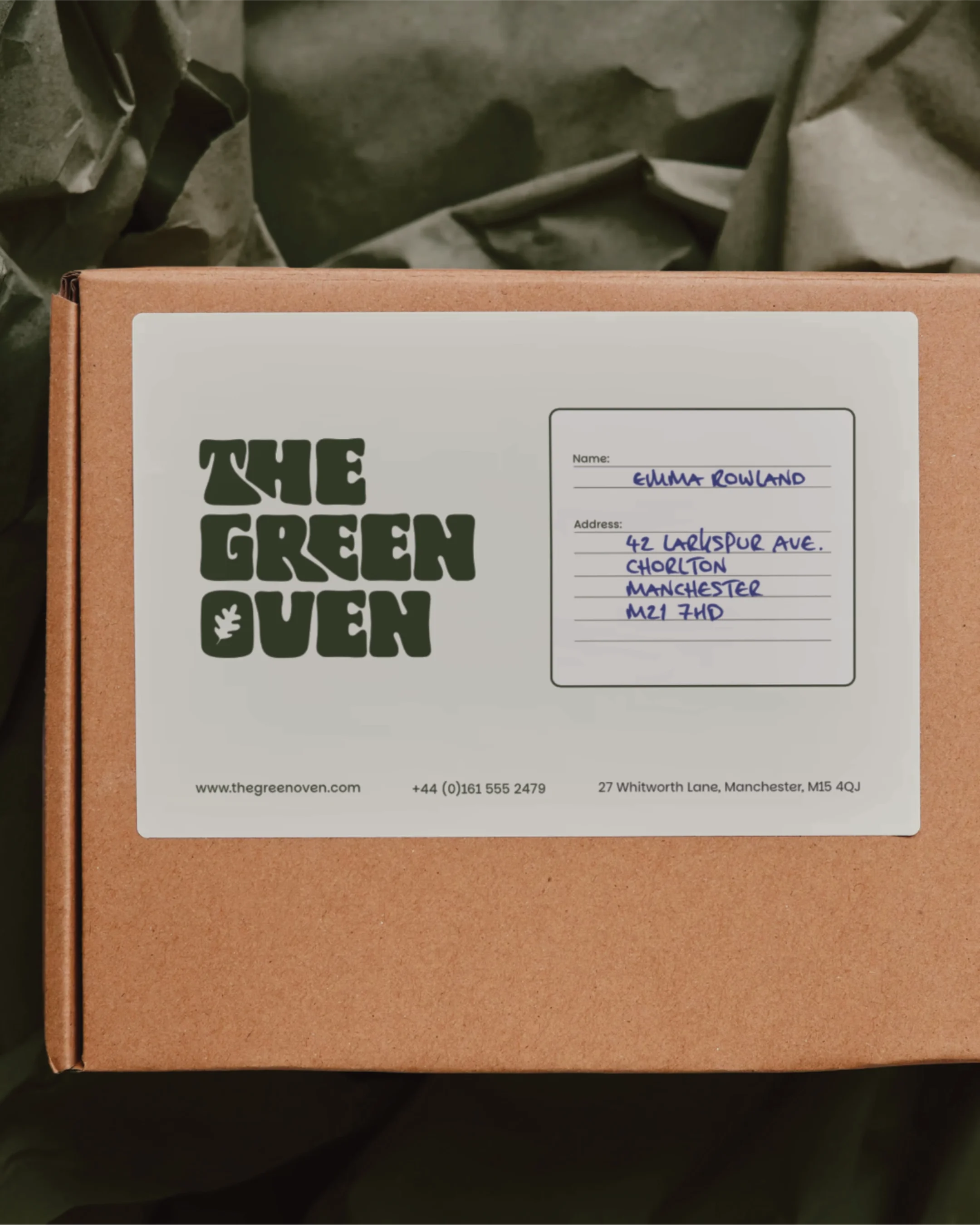 A brown cardboard box with a white label from The Green Oven, featuring the company's logo, website, phone number, and address, along with a handwritten recipient name and address.