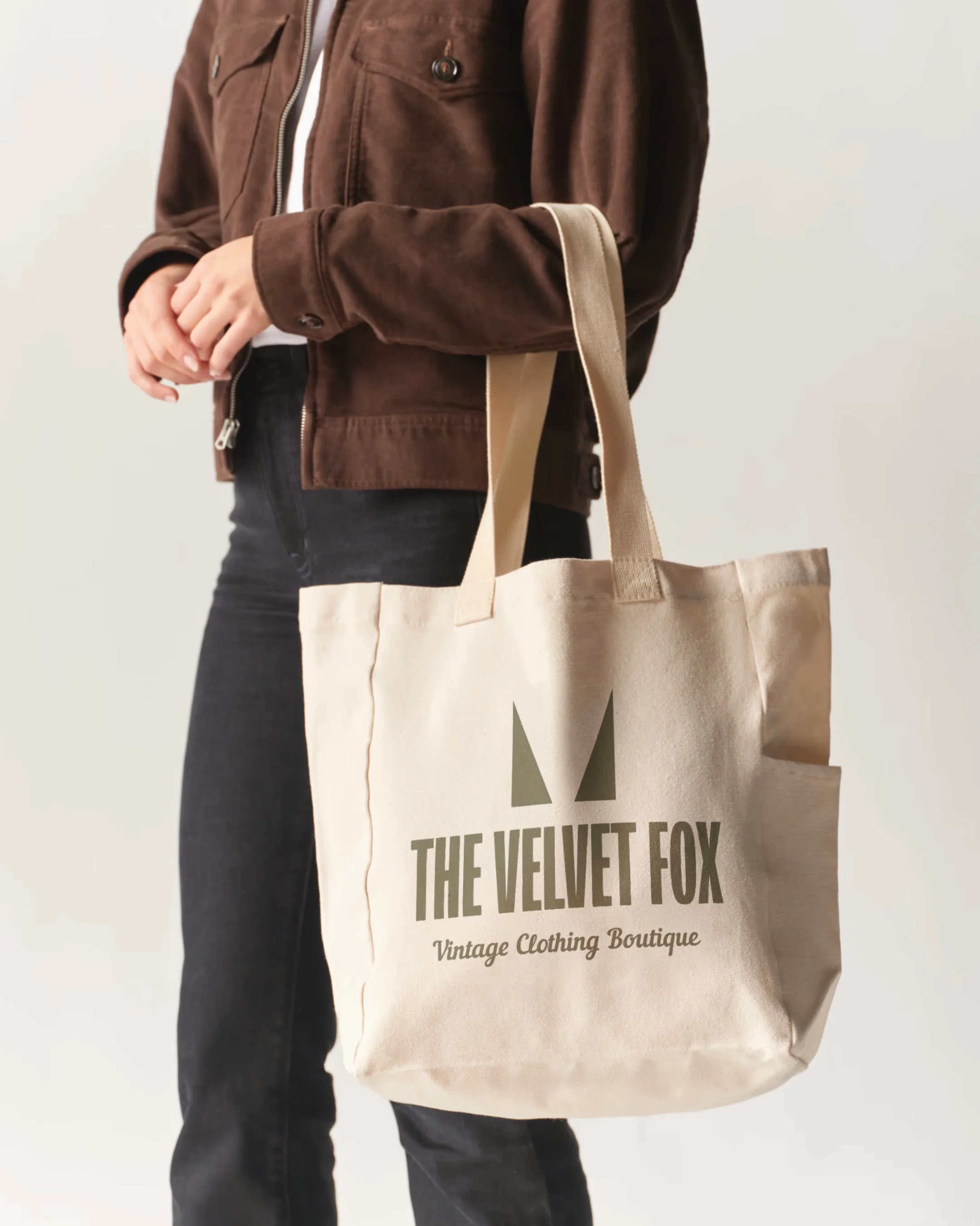 Person holding a beige tote bag with the logo 'The Velvet Fox Vintage Clothing Boutique' printed on it.
