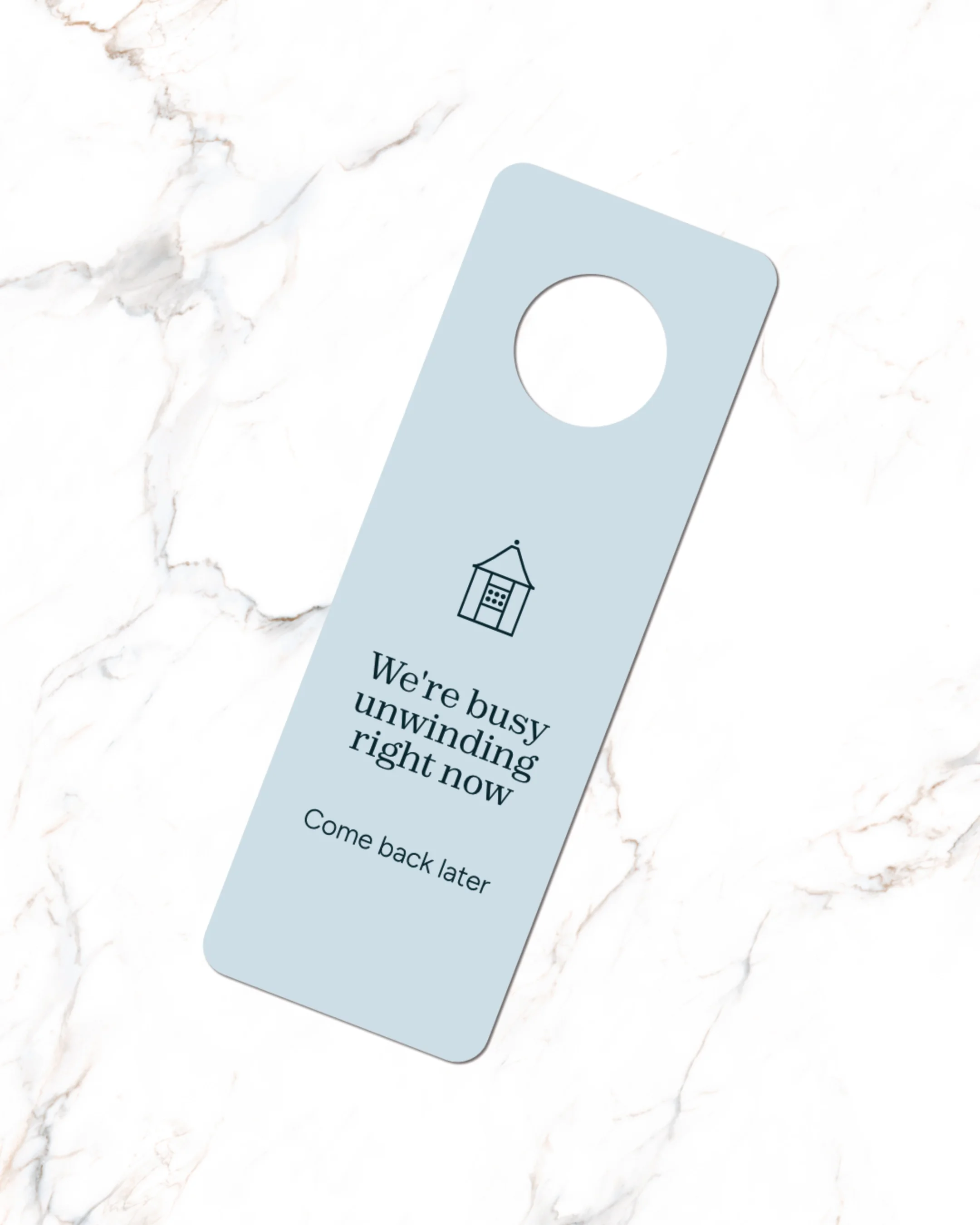 A light blue door hanger with a house icon and the message "We're busy unwinding right now. Come back later" on a white marble surface.