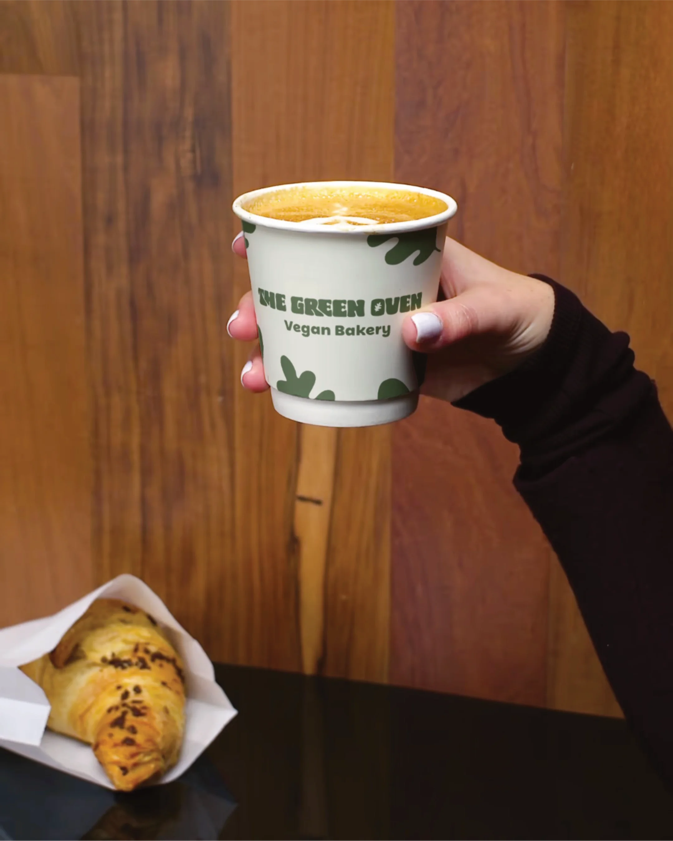 Hand holding a paper cup with the text 'The Green Oven Vegan Bakery' filled with a hot beverage, and a pastry on a black surface against a wooden background.