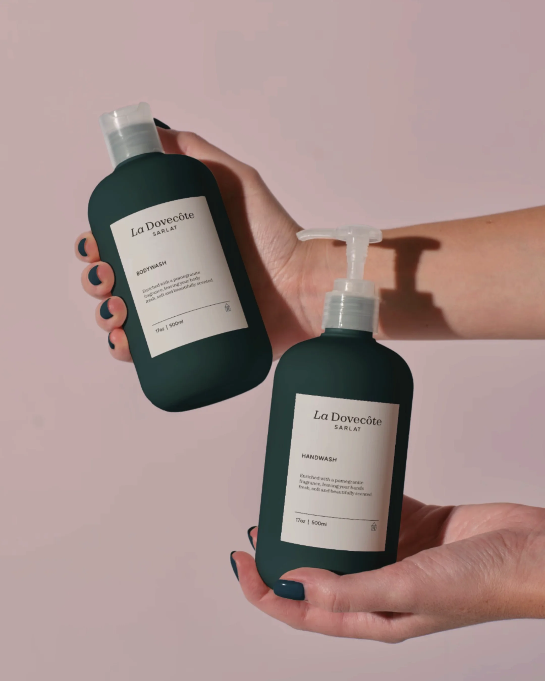 Two hands holding dark green bottles of La Dovecôte Sarlat body wash and hand wash against a pale pink background.