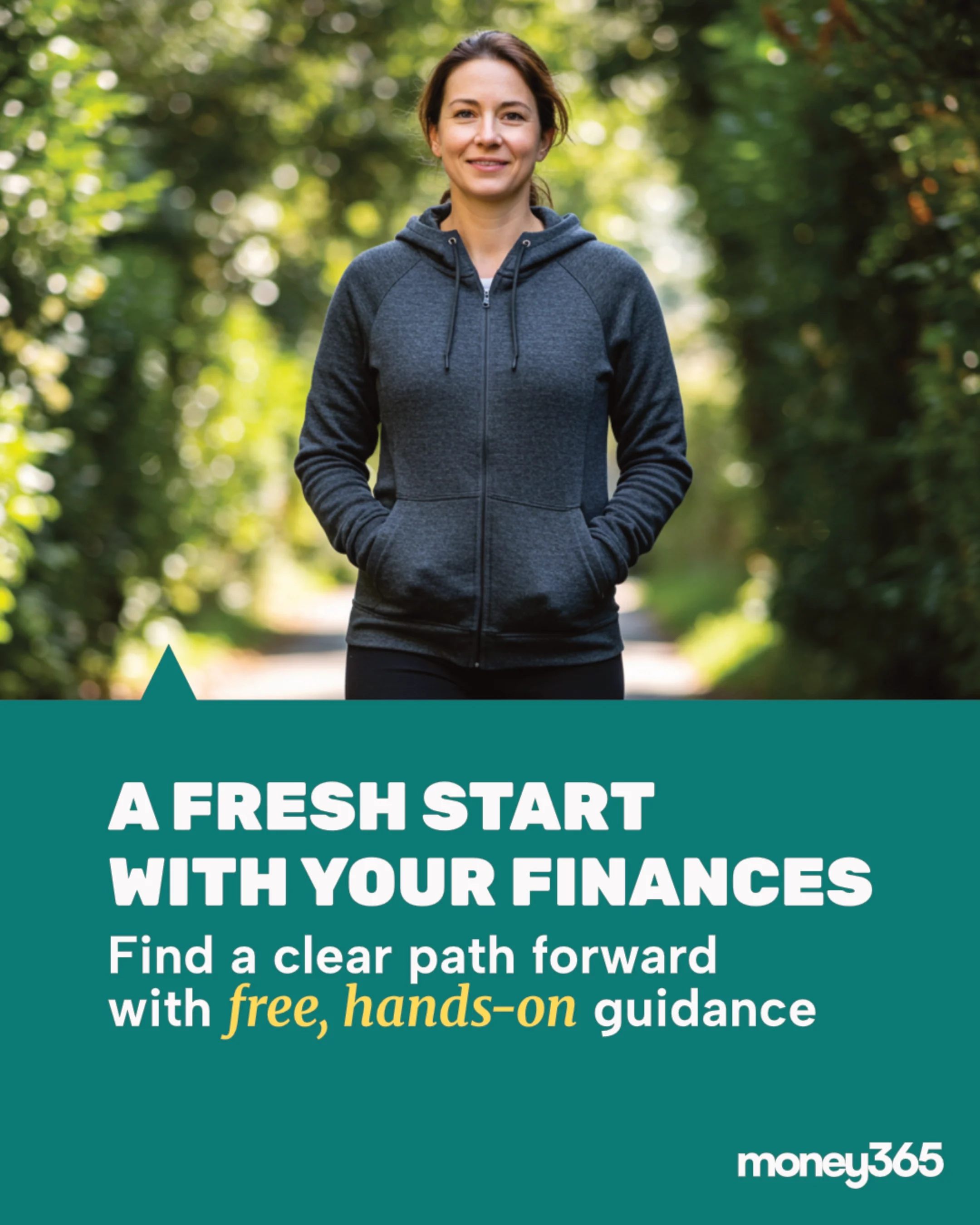 A woman in a dark gray hoodie walking outdoors in a green wooded area, smiling at the camera.