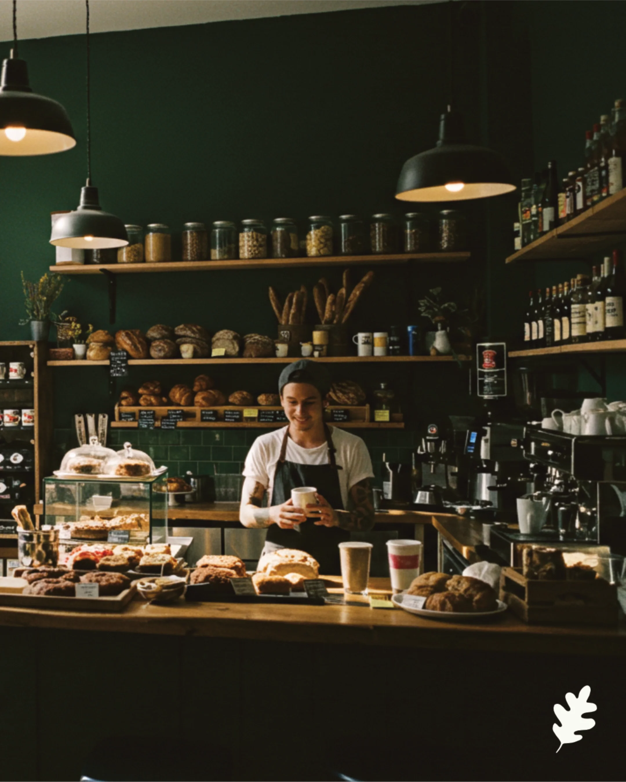 Barista in a coffee shop preparing drinks with baked goods displayed on the counter, shelves of jars and bottles behind her, warm lighting, dark green walls.
