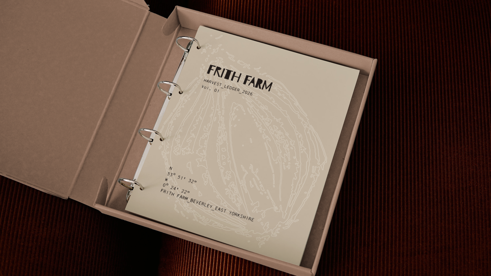 Open cardboard box containing a harvest ledger for Frith Farm, 2026, with anatomical drawings and GPS coordinates on the cover, placed on a dark reddish-brown surface.