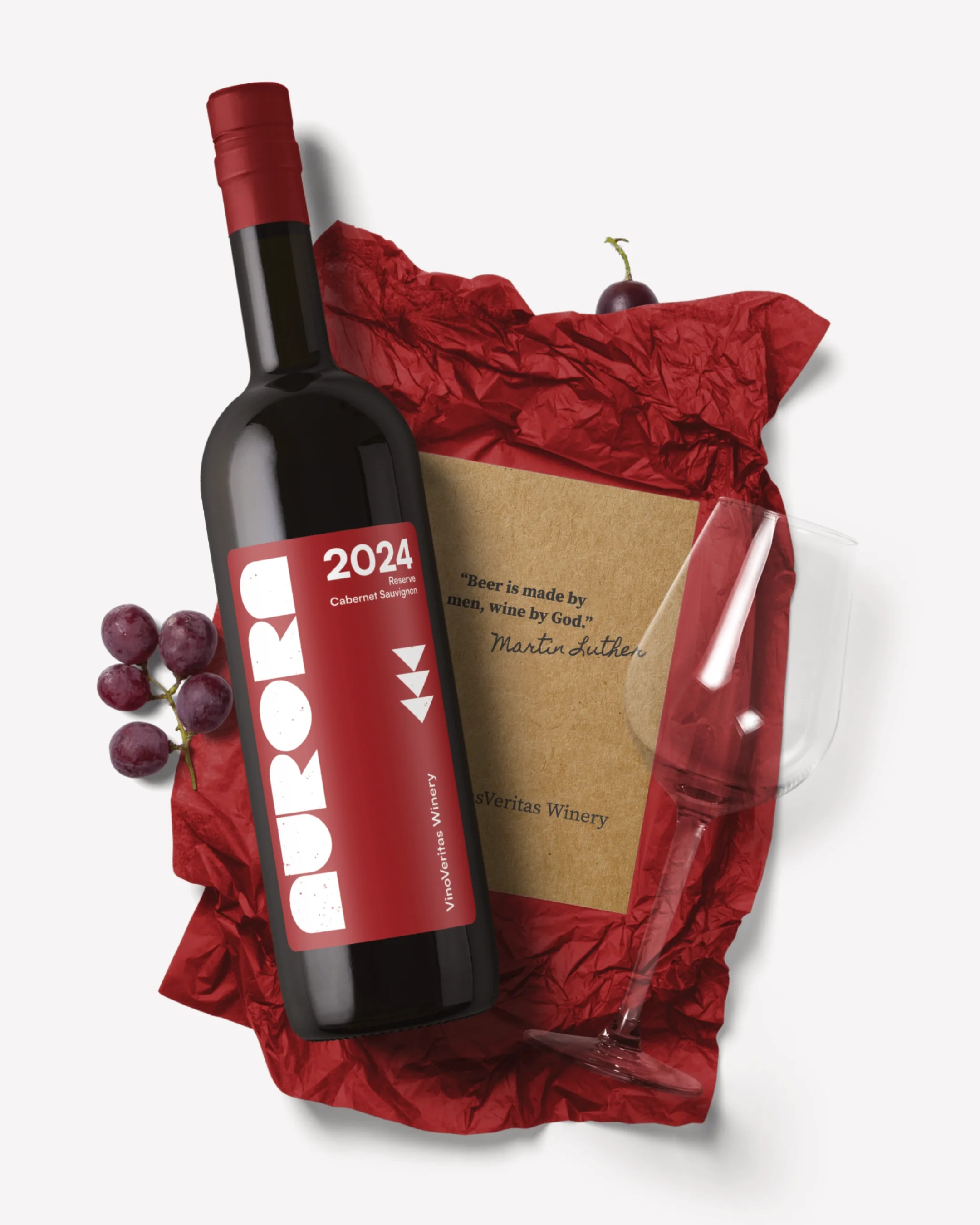 A bottle of red wine, a bunch of grapes, a wine glass, and a gift box with a quote on it, all arranged on red tissue paper.