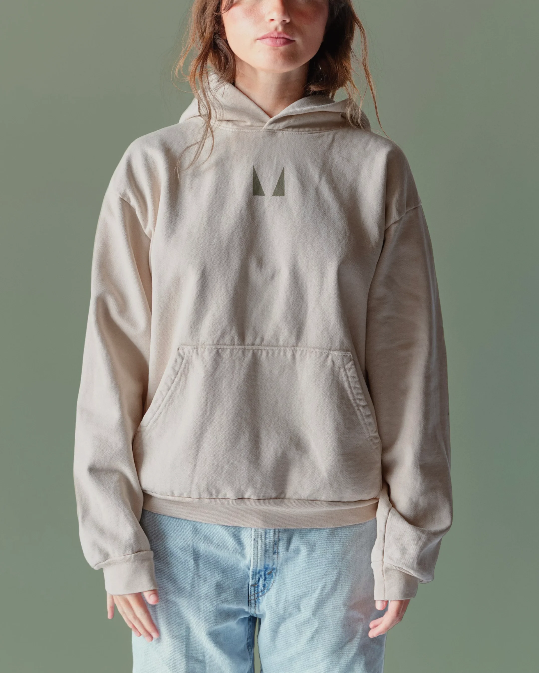 Woman wearing a beige hoodie with a front pouch pocket, light blue jeans, and a small logo on the chest.