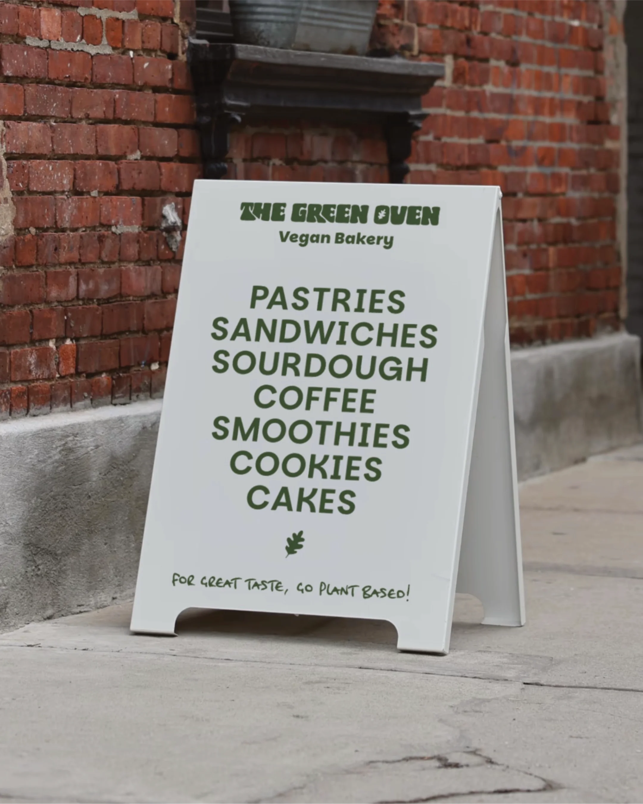 A white A-frame sidewalk sign outside a brick building advertising a vegan bakery called The Green Oven. The sign lists menu items such as pastries, sandwiches, sourdough, coffee, smoothies, cookies, and cakes, with a note promoting plant-based options.