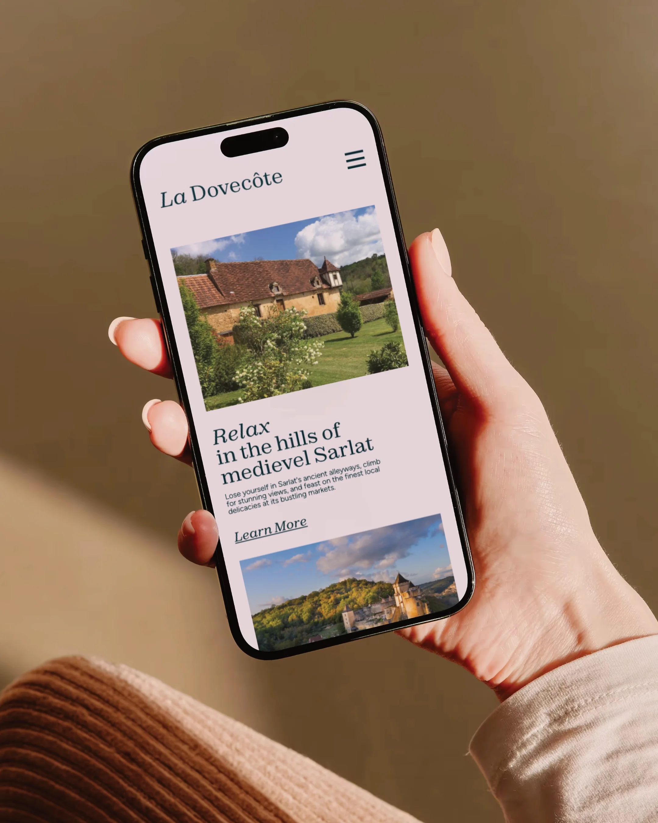 A person holding a smartphone displaying a travel website for La Dovecôte, showing images of a house and scenic landscape with text promoting relaxation in the hills of medieval Sarlat.