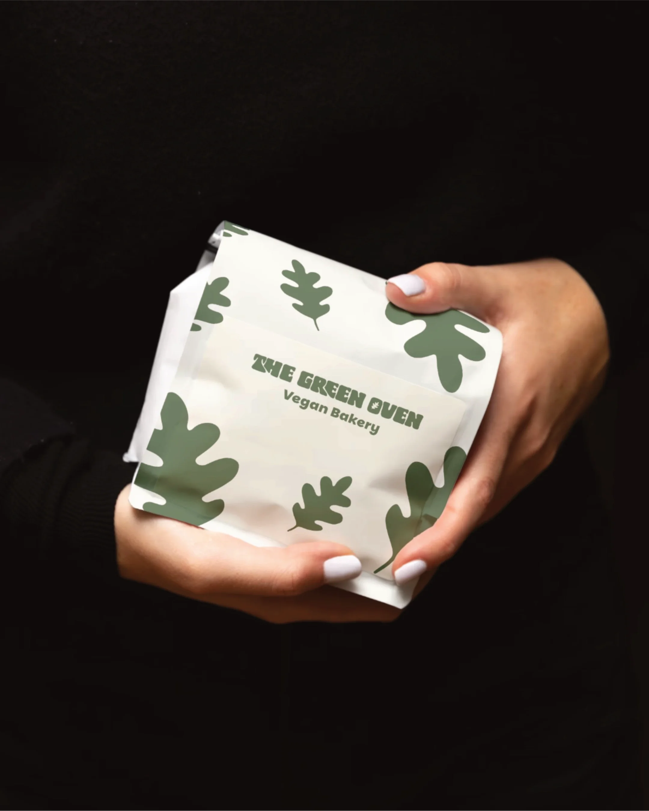 Person holding a white paper bag with green leaf patterns and the text 'The Green Oven Vegan Bakery' printed on it.