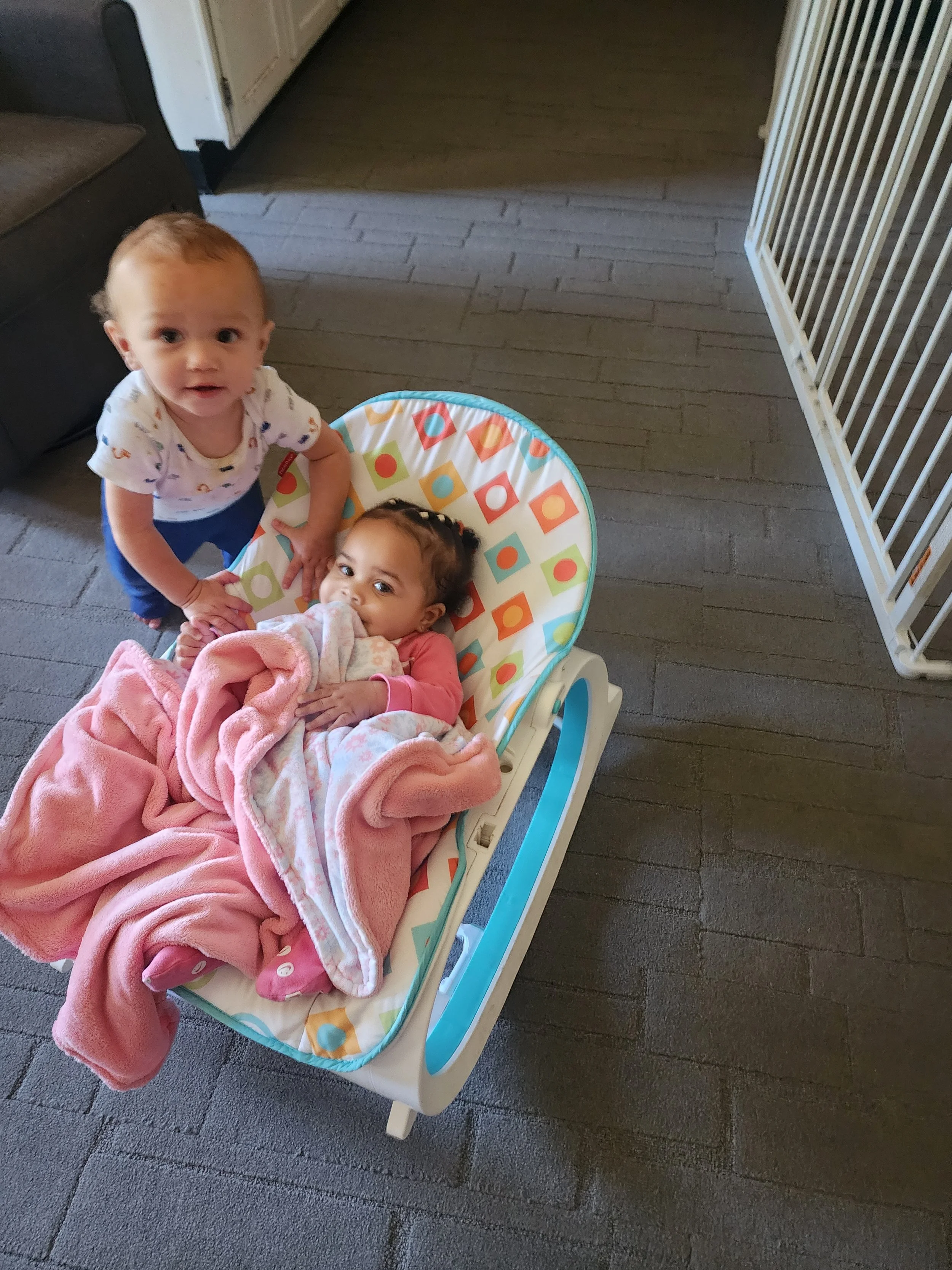 Two children indoors, one standing and the other in a baby seat surrounded by blankets.