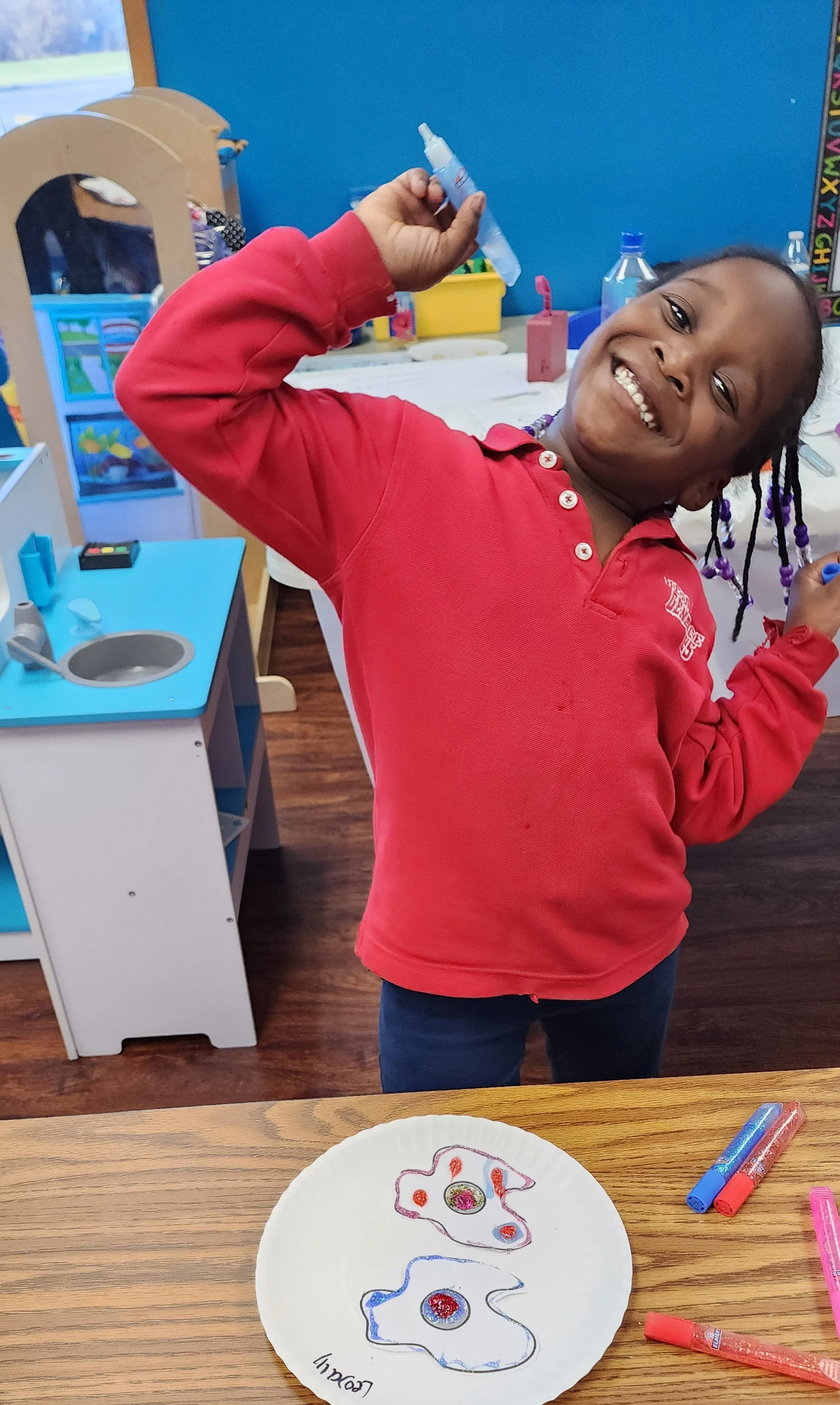 A child in a classroom smiling and holding a marker, with a paper plate featuring colorful designs on a table.