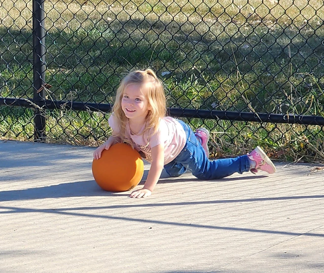 Child on a paved surface playing with an orange ball, with a chain-link fence and grassy area in the background.