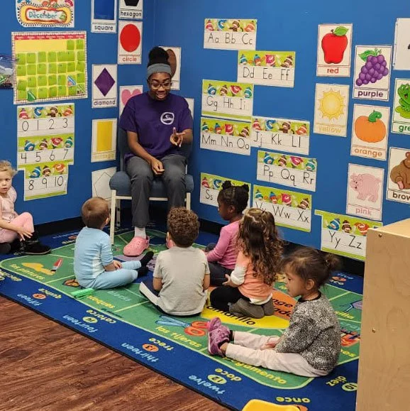 A teacher sitting in a classroom with young children seated around her on a colorful rug, surrounded by educational posters and alphabet charts on the walls.