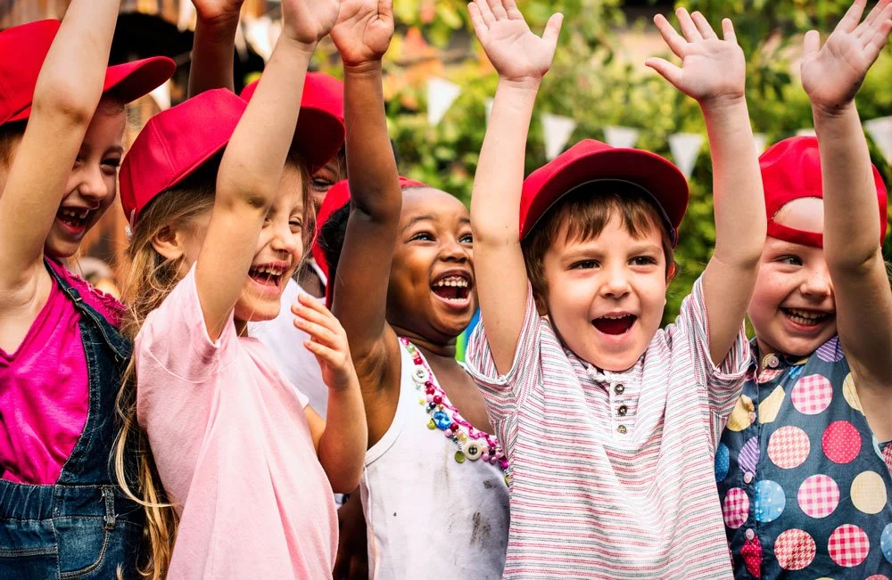 Group of happy children with raised arms, wearing red caps and smiling outdoors.