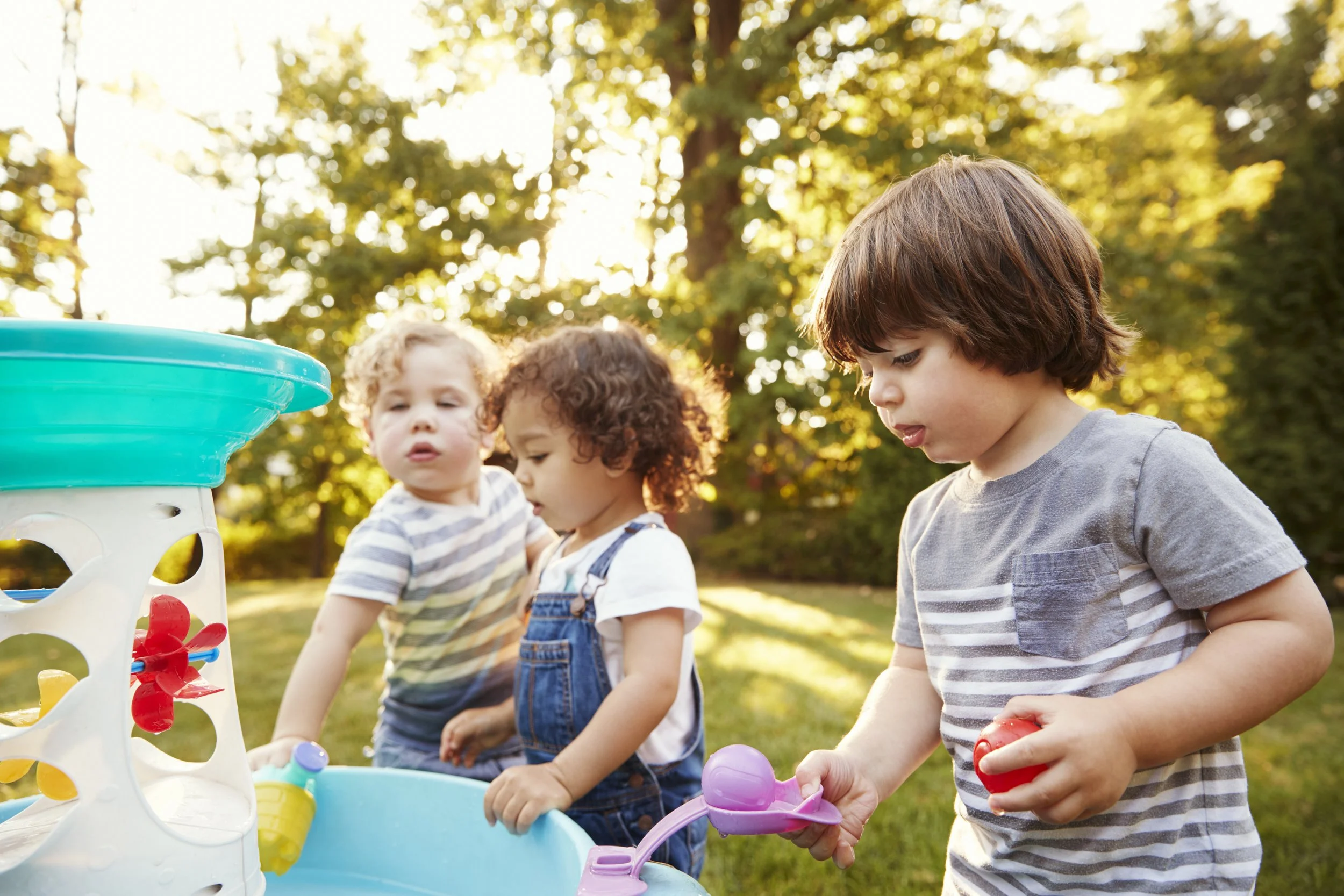 Three young children playing with a water table outdoors in a sunny park.
