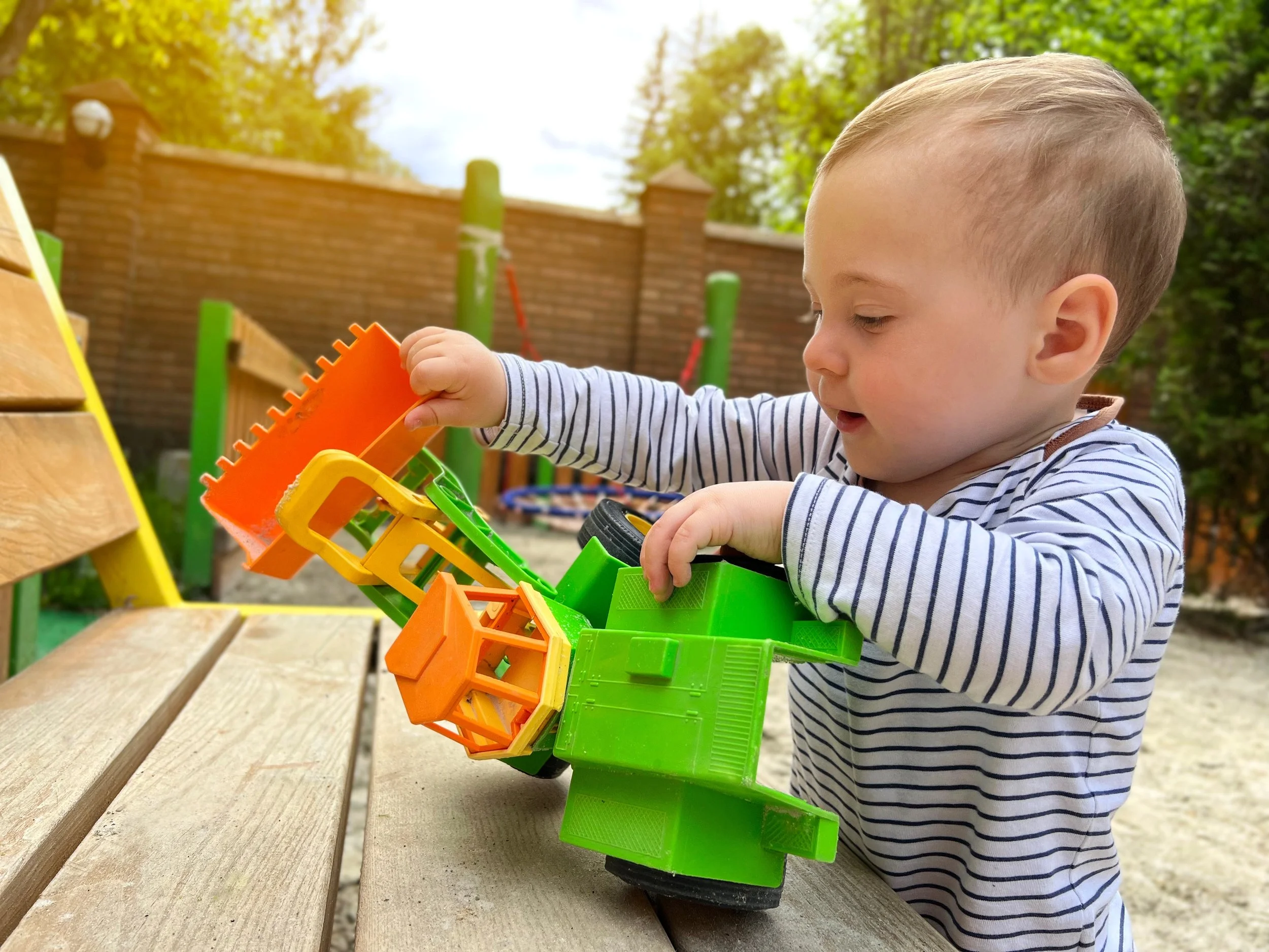 Toddler playing with a green toy tractor on a wooden bench in a park.