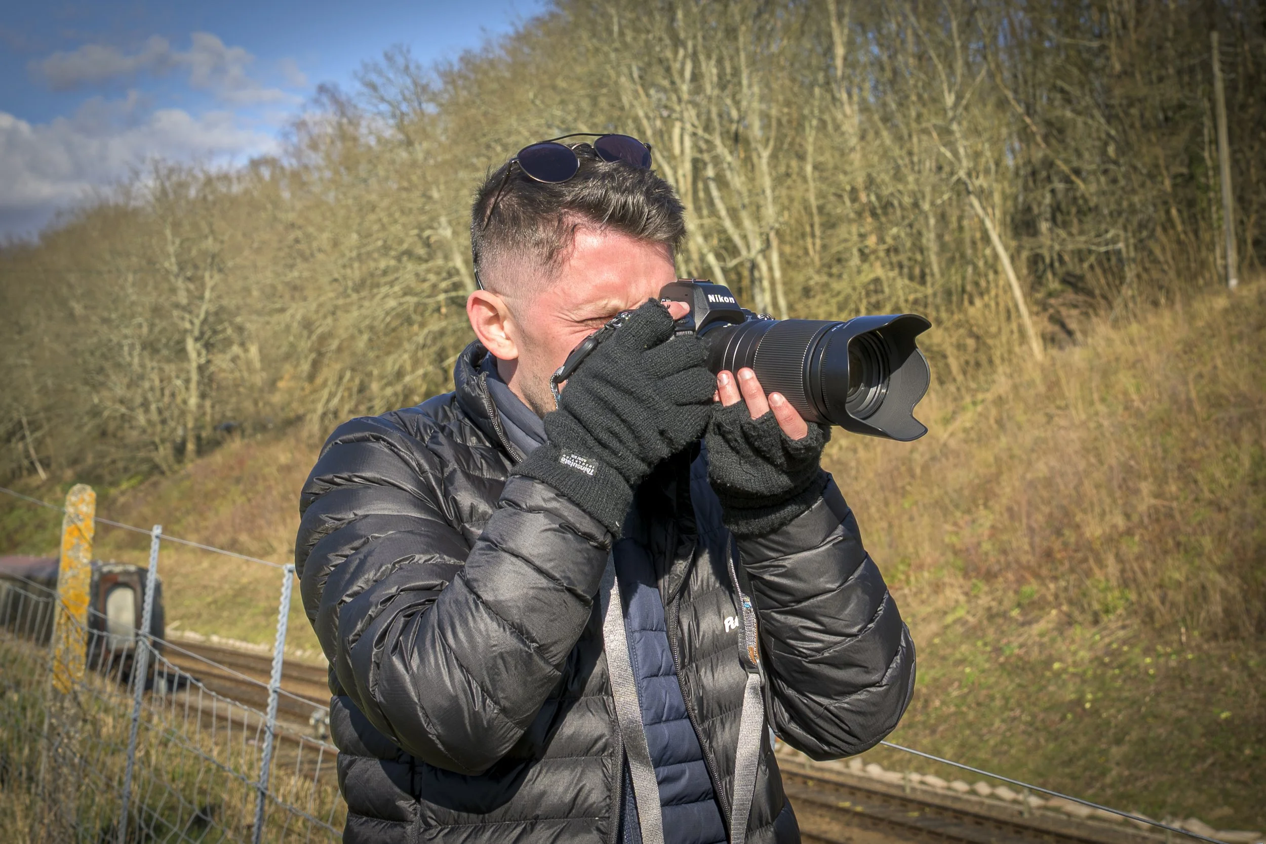 A man with sunglasses on his head, wearing a black jacket and gloves, is taking a photo with a Nikon camera outdoors near a railroad track and trees.