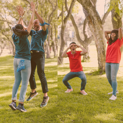 Four friends celebrating and having fun outdoors in a park, with two jumping and high-fiving, one squatting excitedly, and another smiling with her hand on her head.