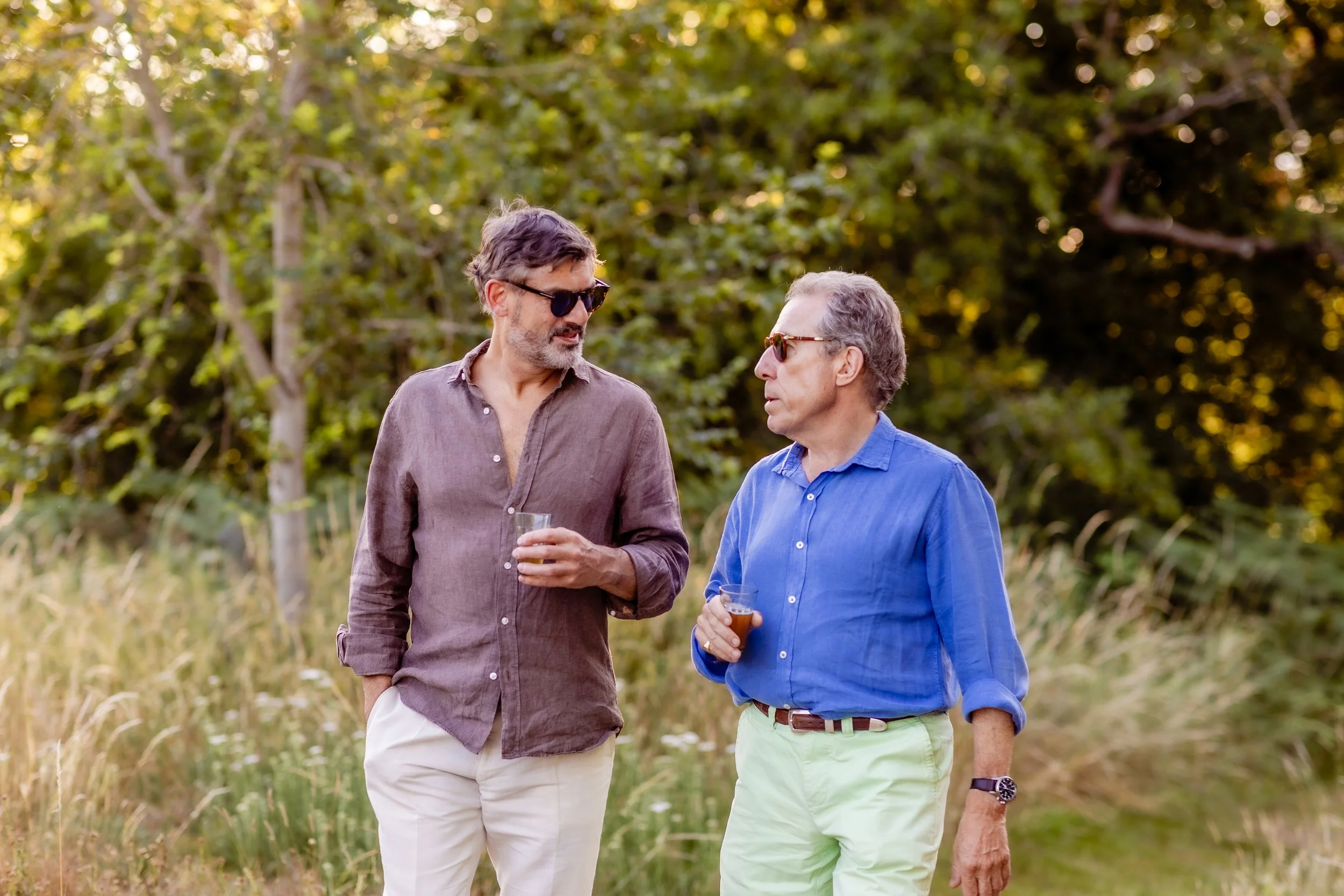 Two men wearing sunglasses and holding drinks, standing in a grassy outdoor area with trees in the background, engaged in conversation.