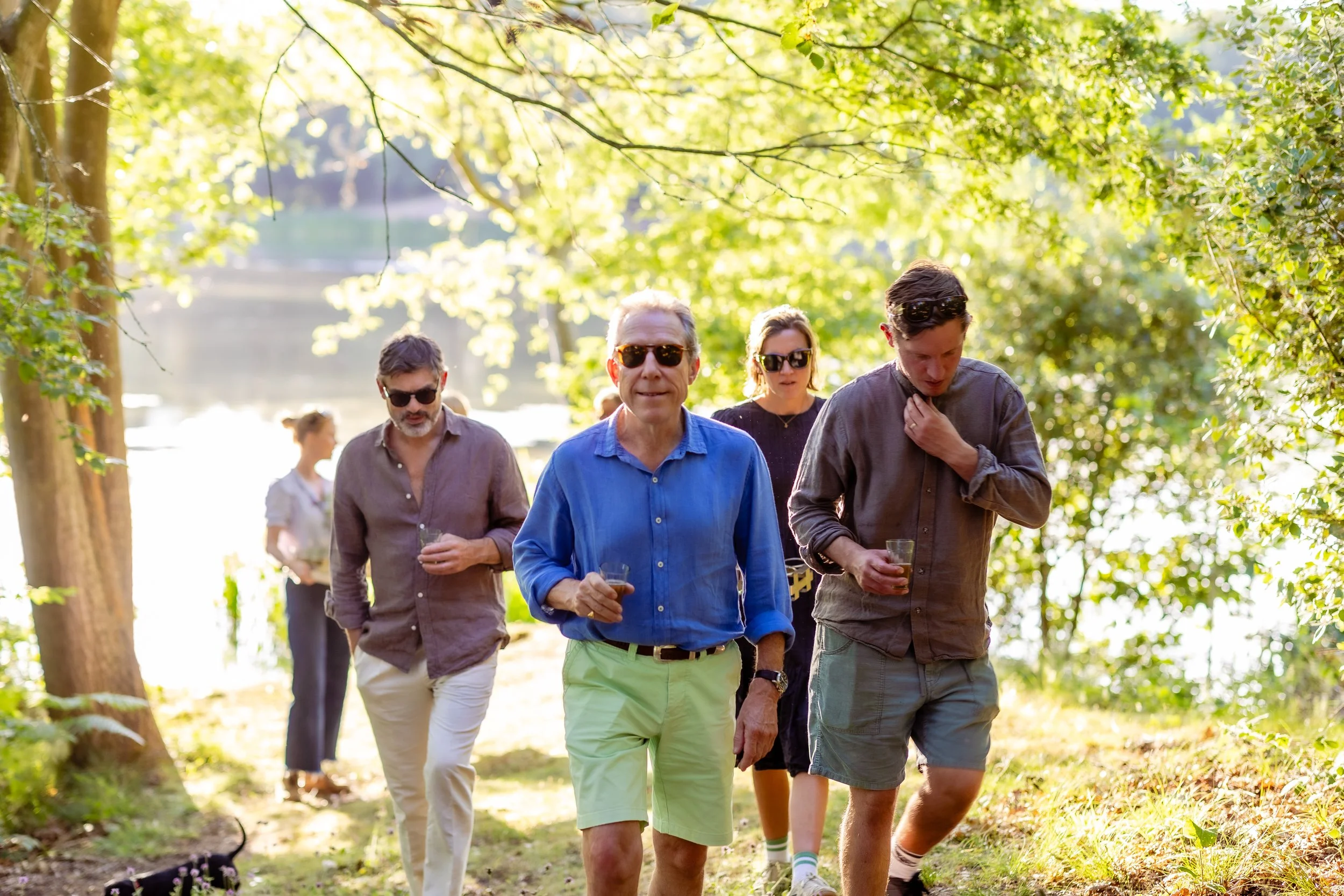 A group of people walking outdoors in a forested area during daylight, some holding drinks and wearing sunglasses.