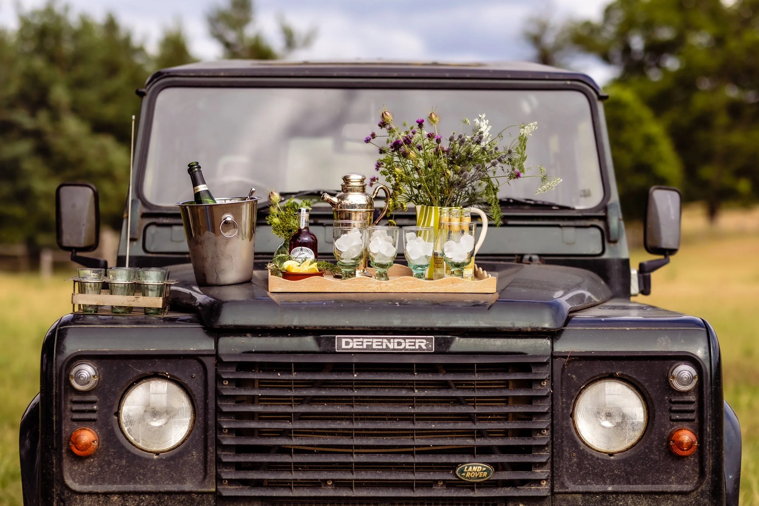 The front of a black Land Rover Defender with a tray on the hood holding glasses of ice, flowers, a teapot, a bottle of syrup, and a champagne bottle in a cooler, against a background of trees and a cloudy sky.