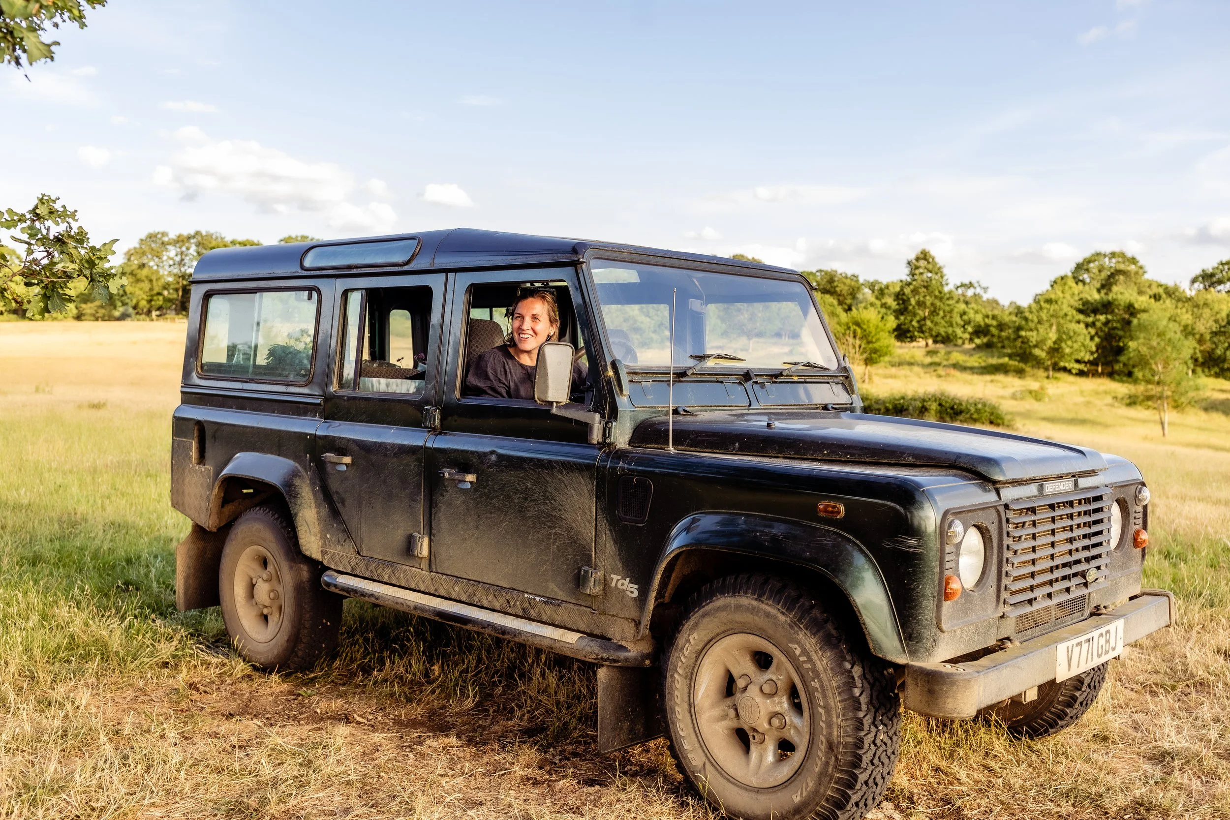 A woman with brown hair sitting in the driver's seat of a black Land Rover Defender vehicle, parked on a grassy field under a partly cloudy sky with trees in the background.
