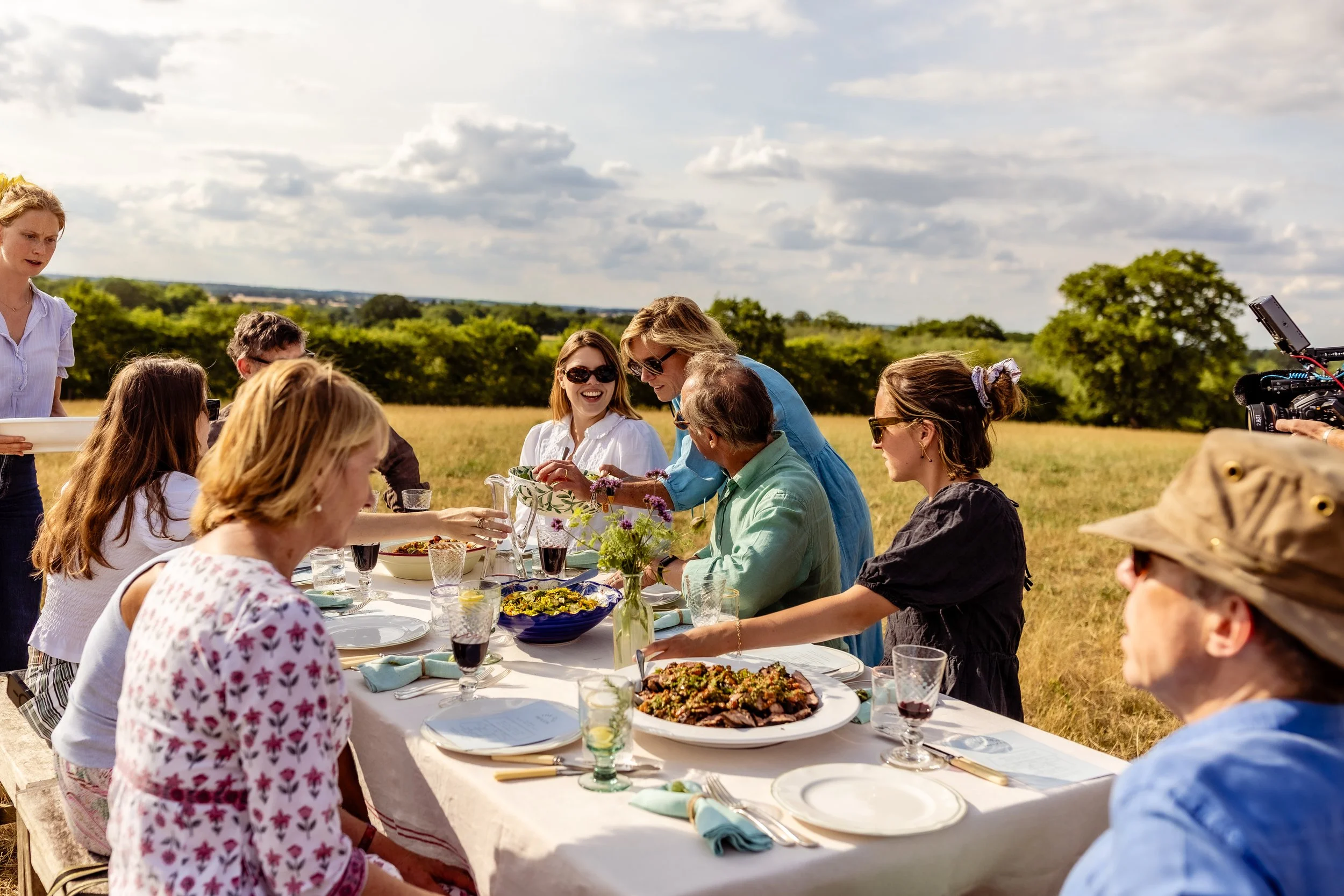 A group of people gathered outdoors having a meal at a long table on a sunny day, with food, drinks, and floral decorations visible.