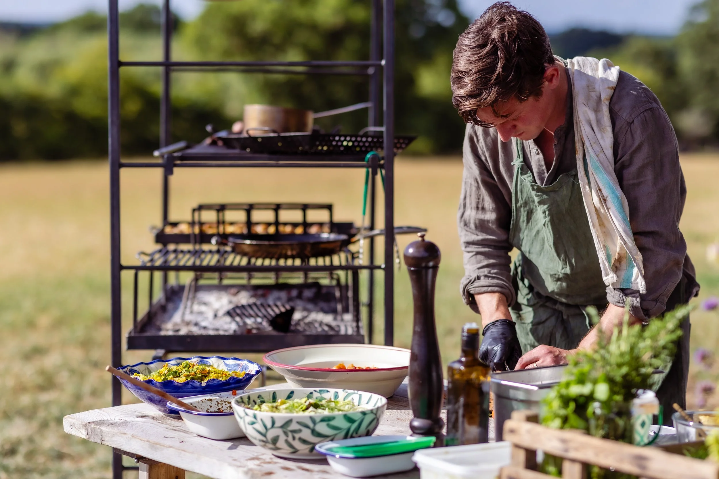 A person preparing food outdoors at a picnic table with bowls of salad and various cooking utensils, while a grill in the background is being used.