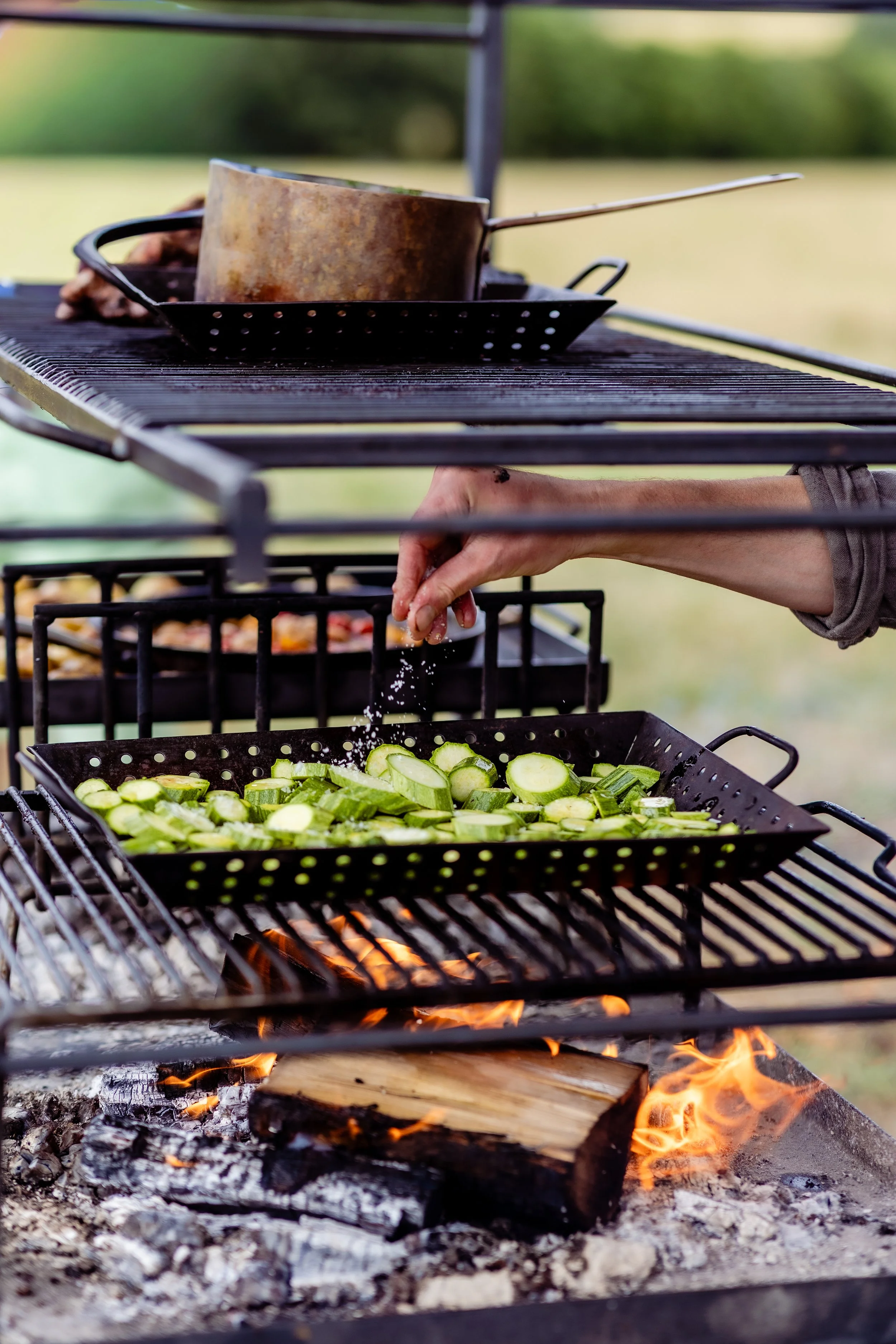 A close-up of a barbecue grill with flaming charcoal, a rack of sliced zucchini, and a person’s hand sprinkling salt on the vegetables. A roasting pan with a copper pot and tongs is on the grill, with a blurred outdoor background.
