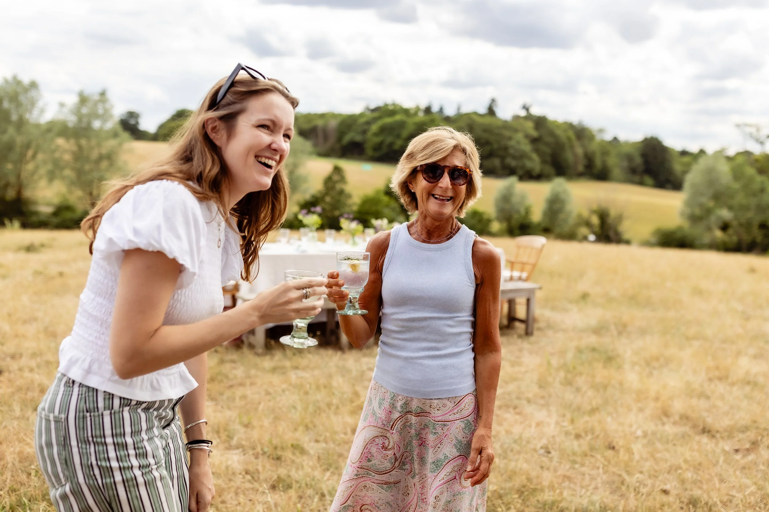 Two women, one young and one older, smiling and holding glasses of drinks outdoors in a field with trees and cloudy sky in the background.