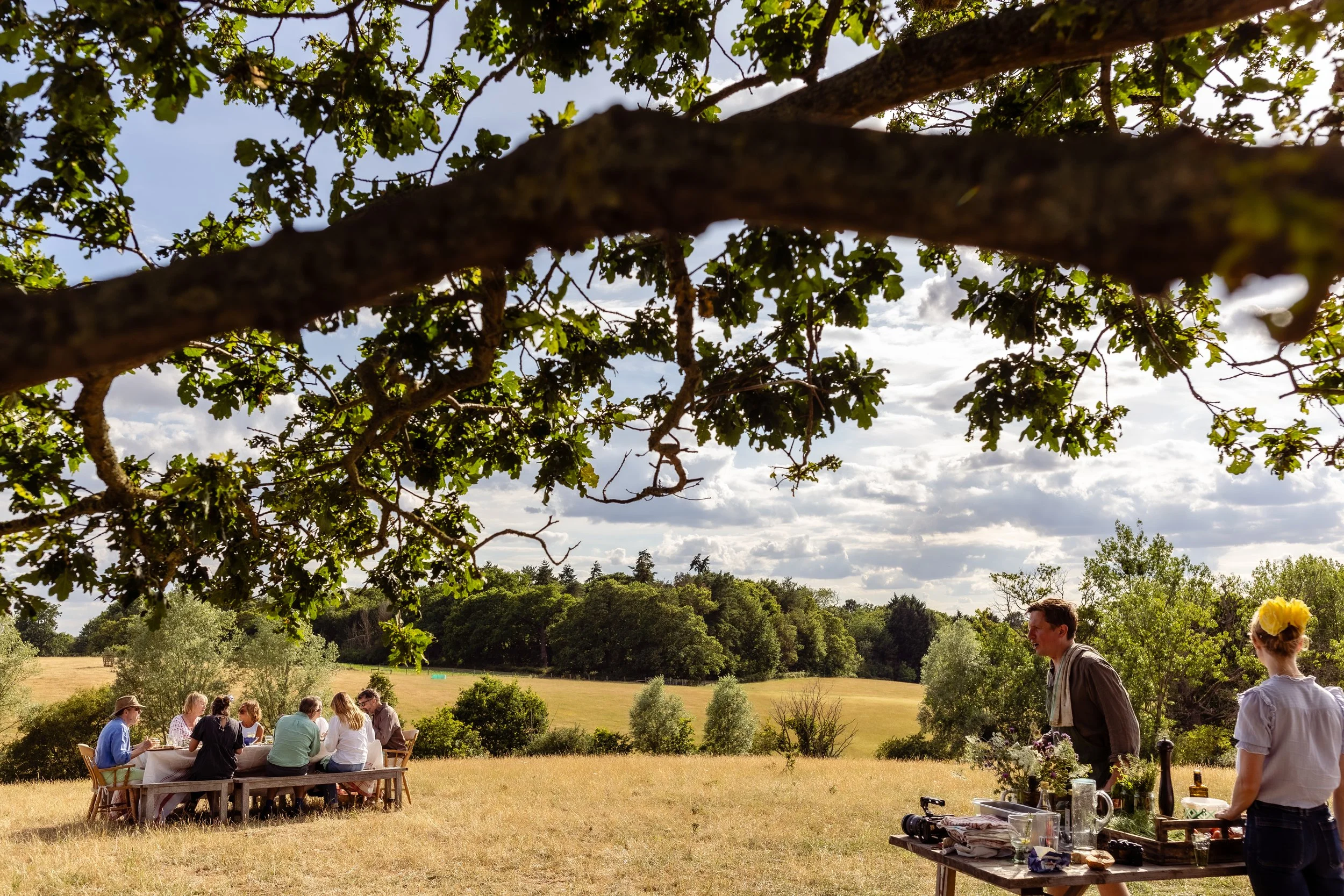 People gathered around a table outdoors under trees with a wide landscape in the background on a partly cloudy day.