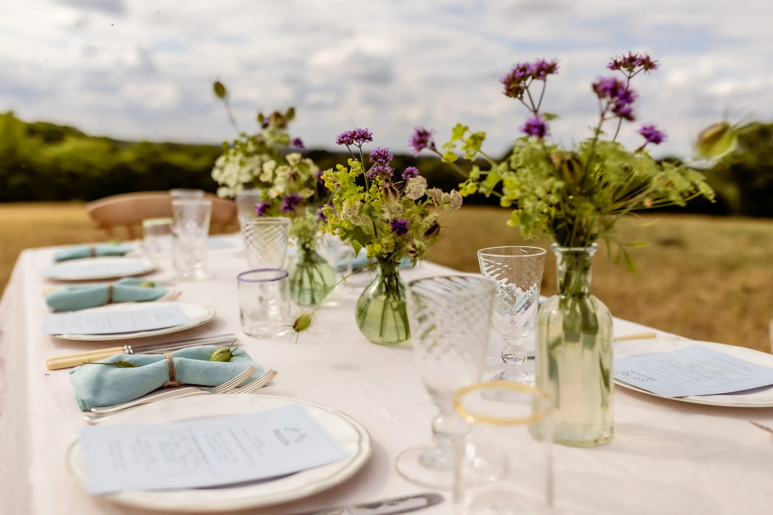 Table set for outdoor event with vases of purple and white flowers, glassware, menus, and light blue napkins, against a background of cloudy sky and open field.