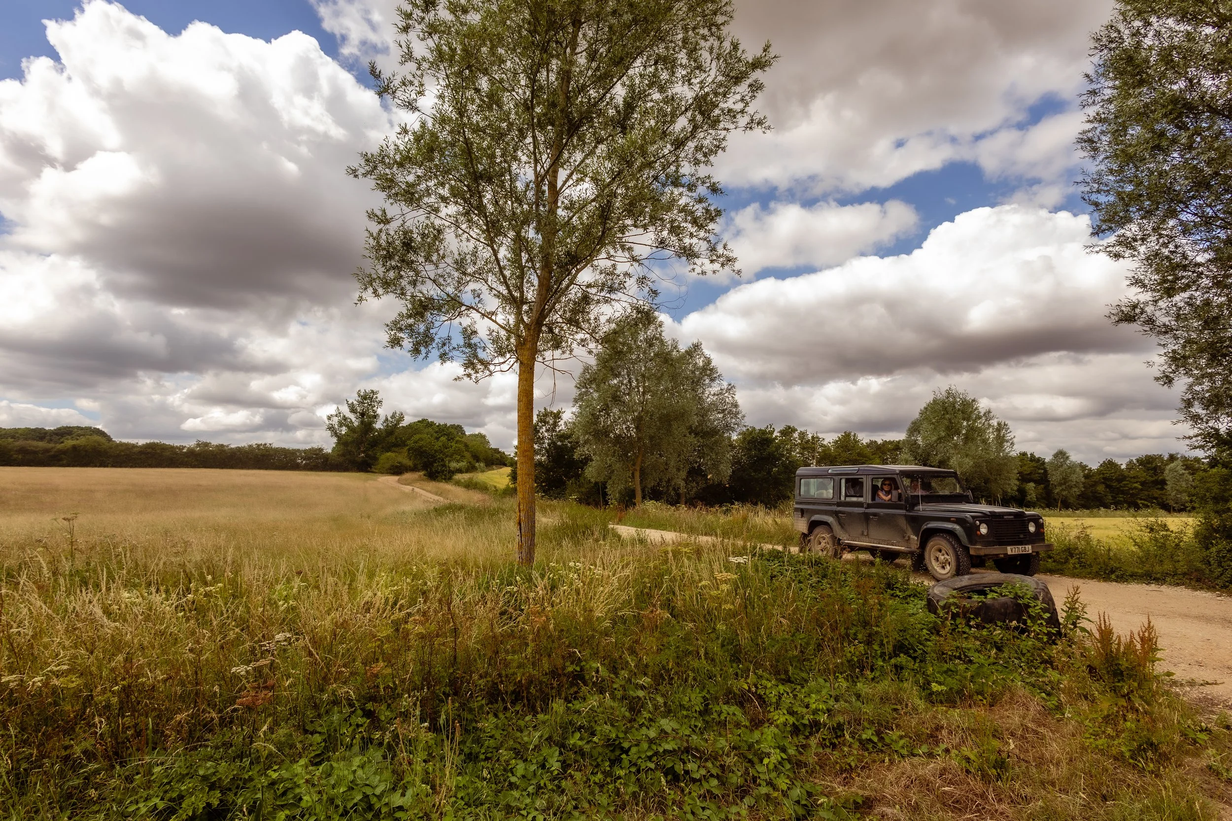 A black off-road vehicle parked on a dirt path in a grassy field with trees and a cloudy sky overhead.