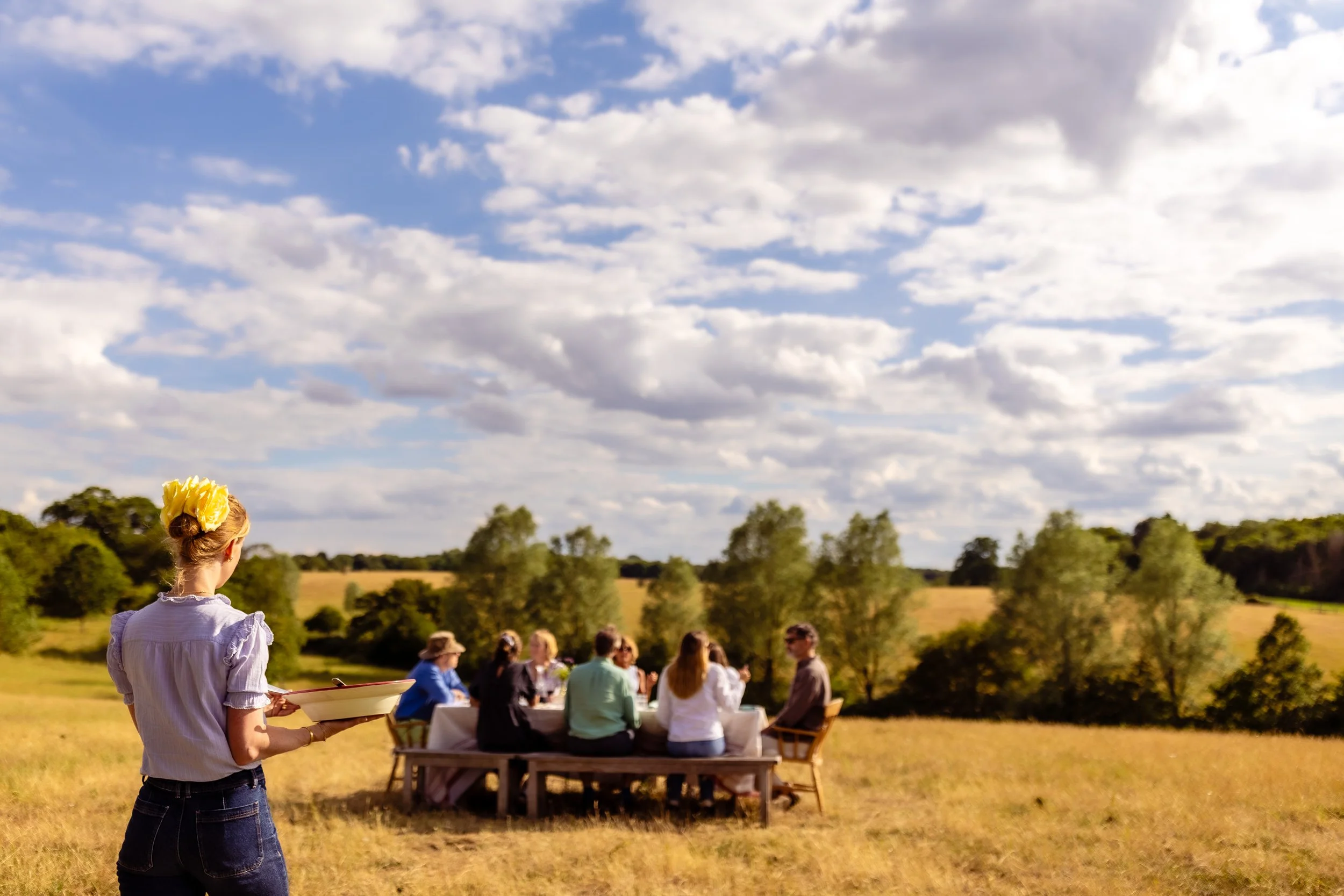 A woman with a yellow flower in her hair, holding a tray with food, stands in front of a group of people sitting at a table outdoors on a grassy field, under a partly cloudy sky.
