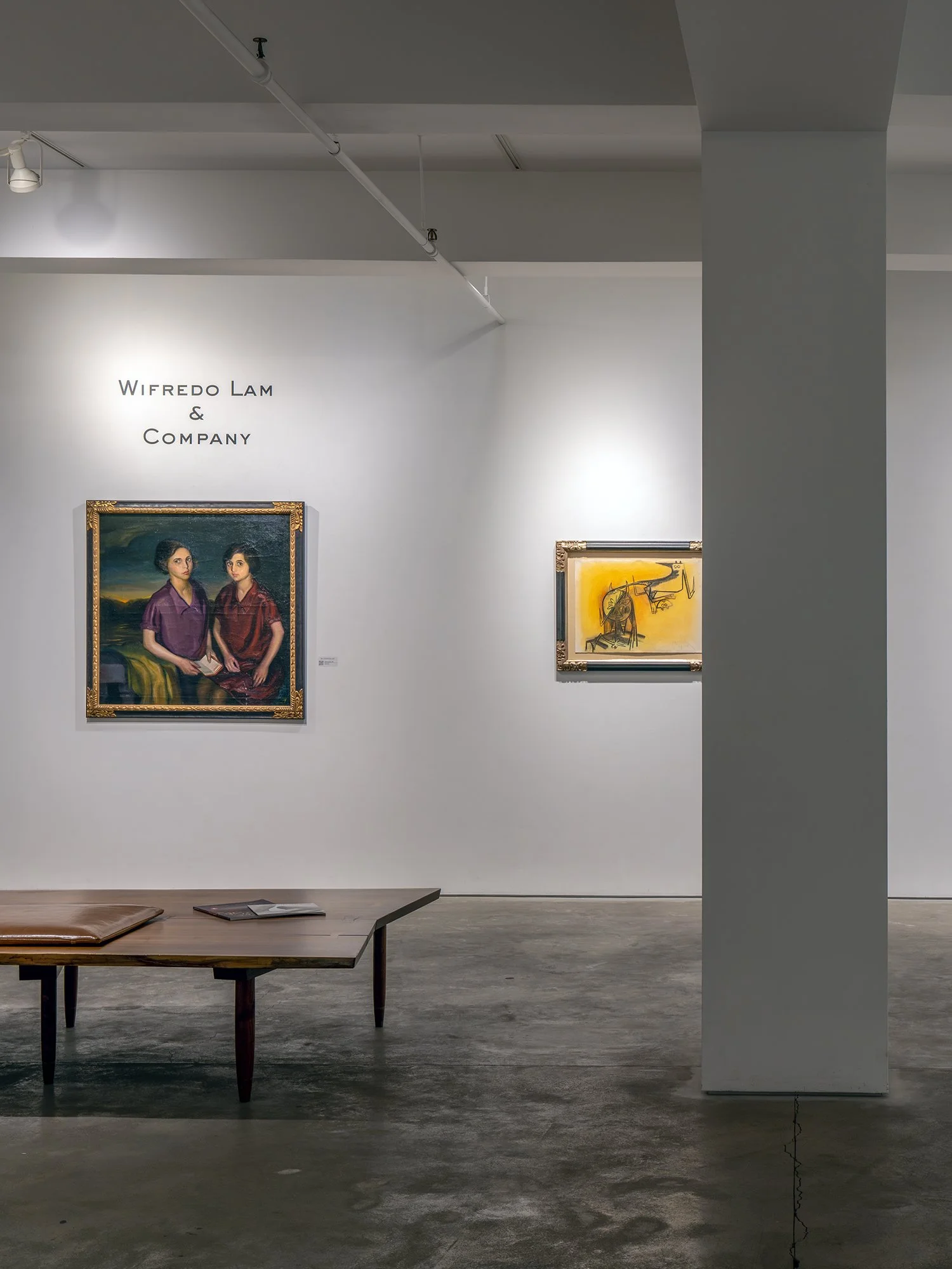 Art gallery with paintings on the wall, wooden table with a cushion and magazines, and the text 'Wifredo Lam & Company' on the wall.