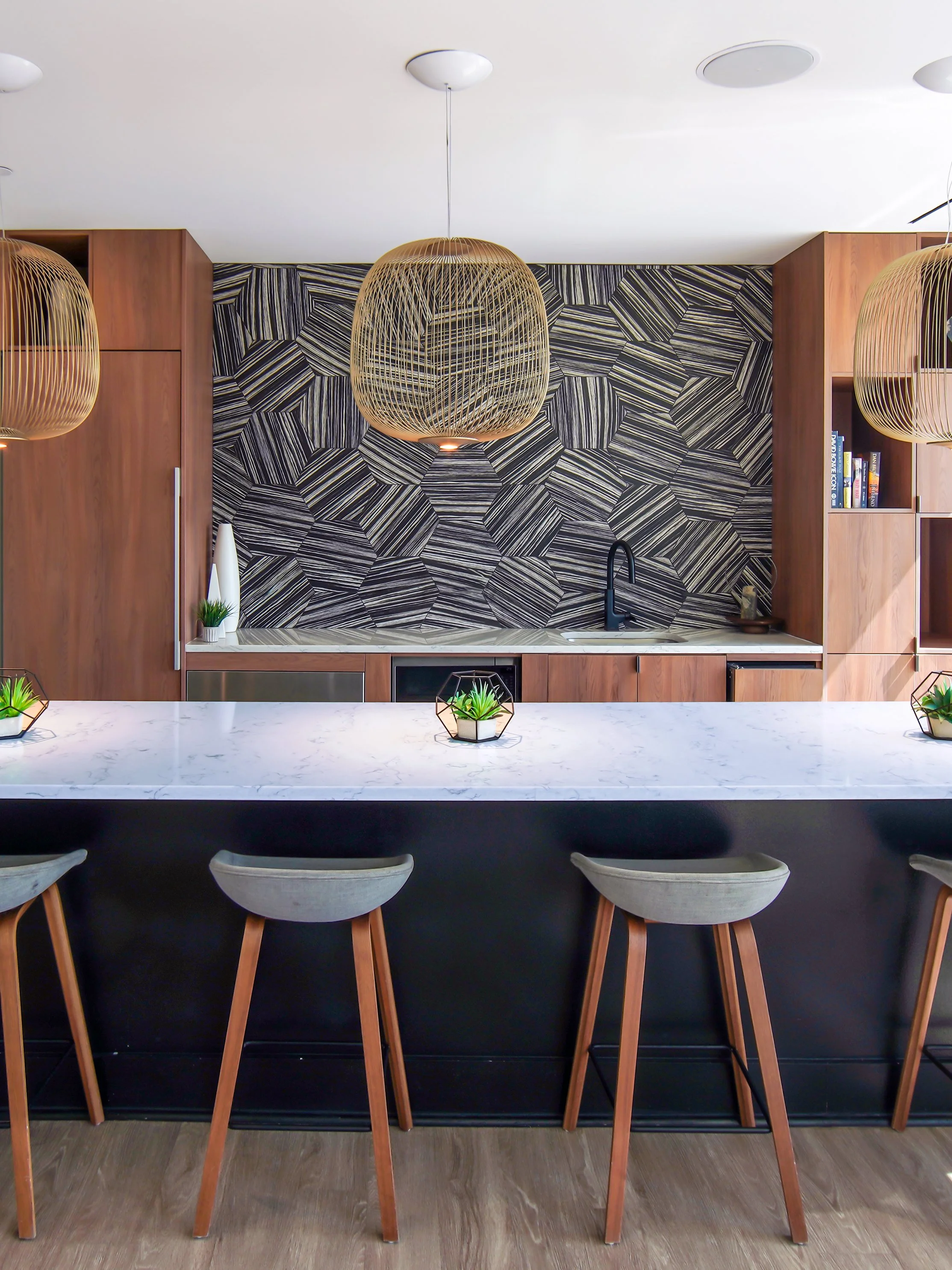 Modern kitchen with patterned black and white backsplash, wooden cabinetry, and a white marble island with three barstools. Hanging wicker pendant lights overhead and small potted plants on the island.