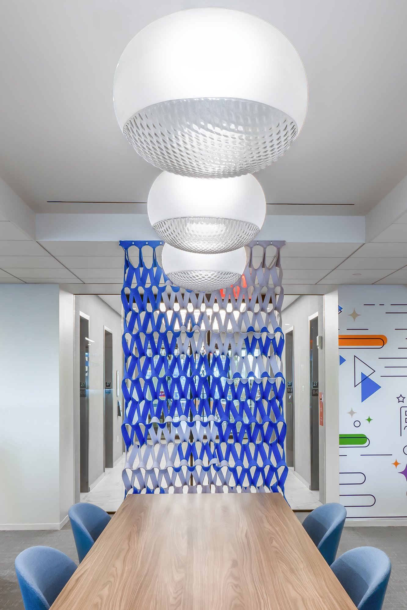 Modern workspace with a wooden table, blue chairs, decorative hanging lights, and an intricate blue and white partition.