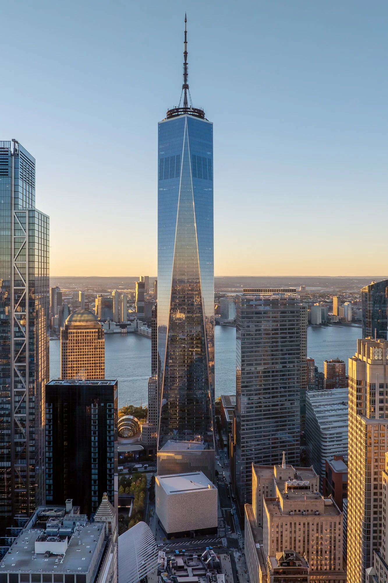 Aerial view of New York City skyline at sunset, featuring One World Trade Center with its reflective glass facade and spire, surrounded by other skyscrapers and the Hudson River in the background.