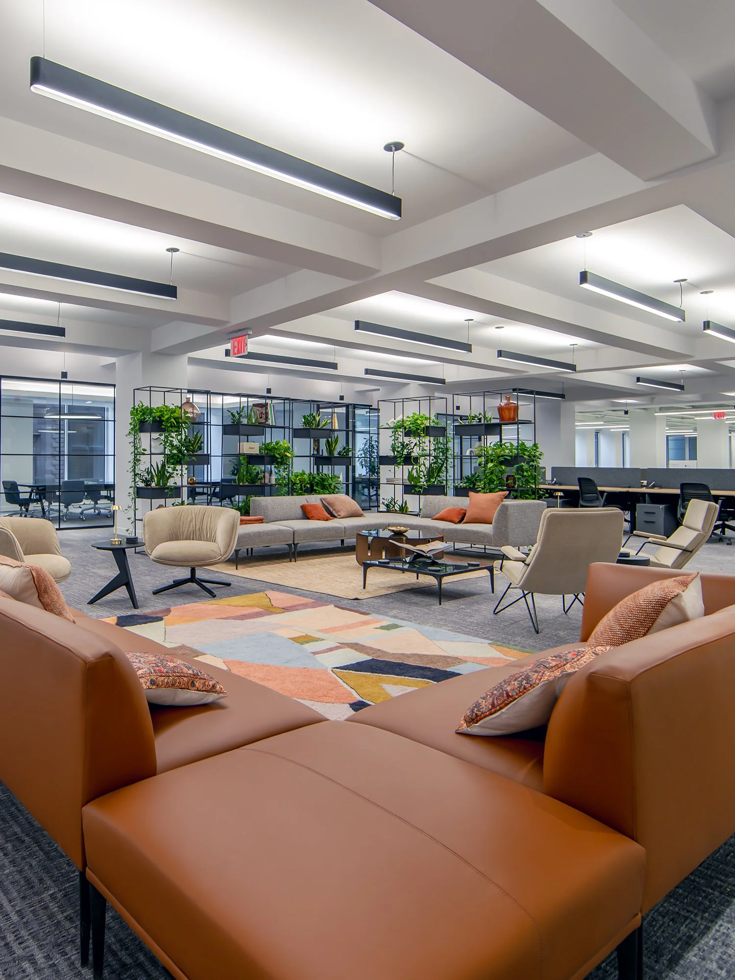 Modern office lounge area with sectional sofa, armchairs, coffee tables, and a multicolored rug, surrounded by bookshelves filled with green plants and decorative items.