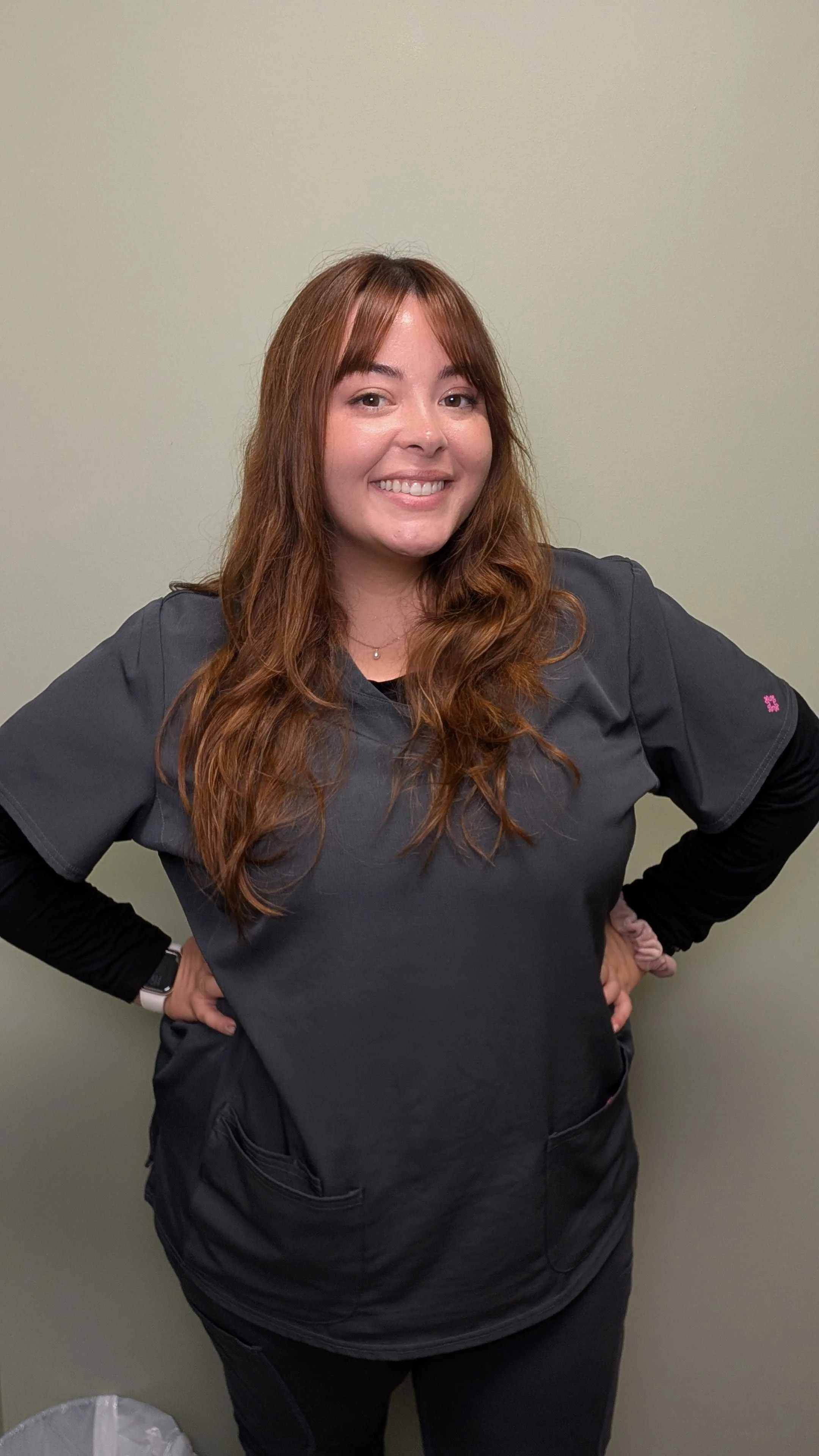 A woman with brown hair tied back, wearing hoop earrings and a dark shirt, smiling in front of a wooden background.