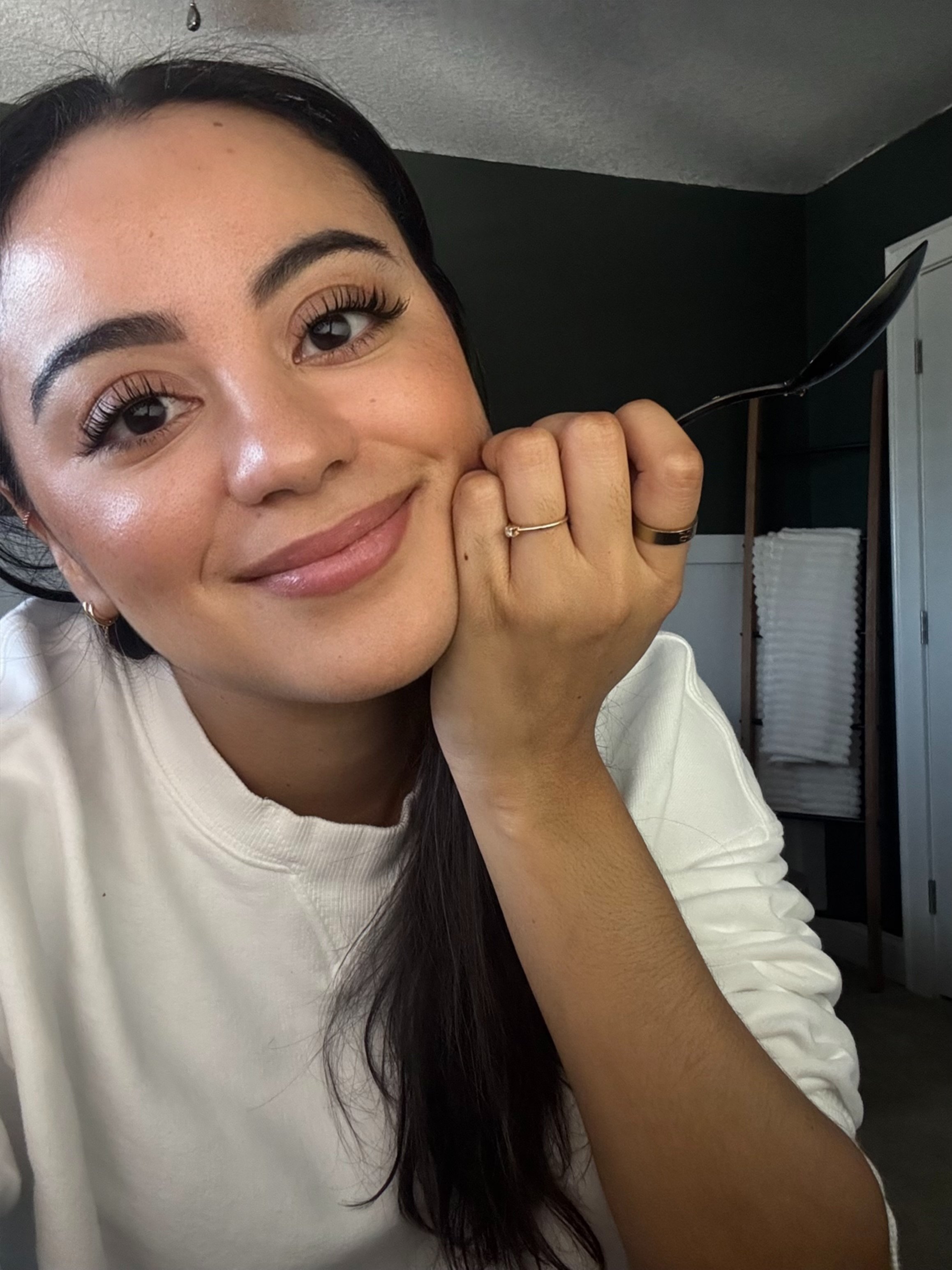 A woman with dark hair and a white shirt smiling, resting her head on her hand, holding a spoon.