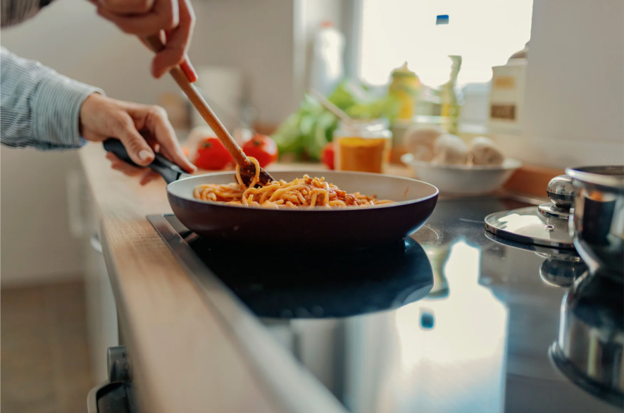 Person cooking spaghetti in a frying pan on stovetop with kitchen ingredients in background.