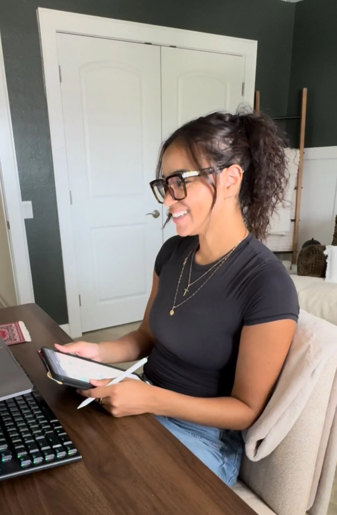 A woman with curly hair, glasses, and wearing earrings and necklaces, sitting at a wooden table, smiling while hosting a telehealth visit.