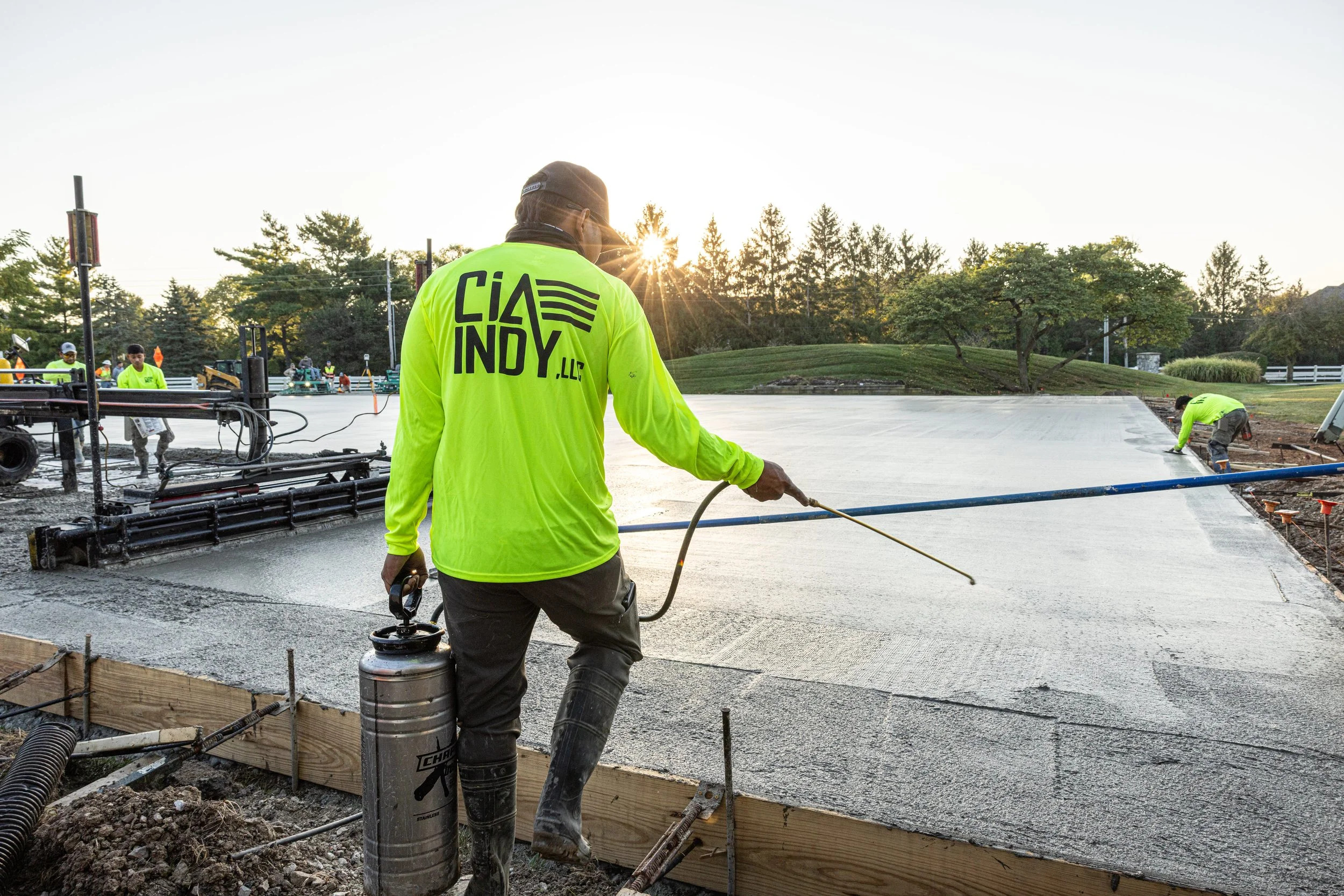 Construction workers pouring and leveling concrete on a building site during sunset, with trees and a white fence in the background.