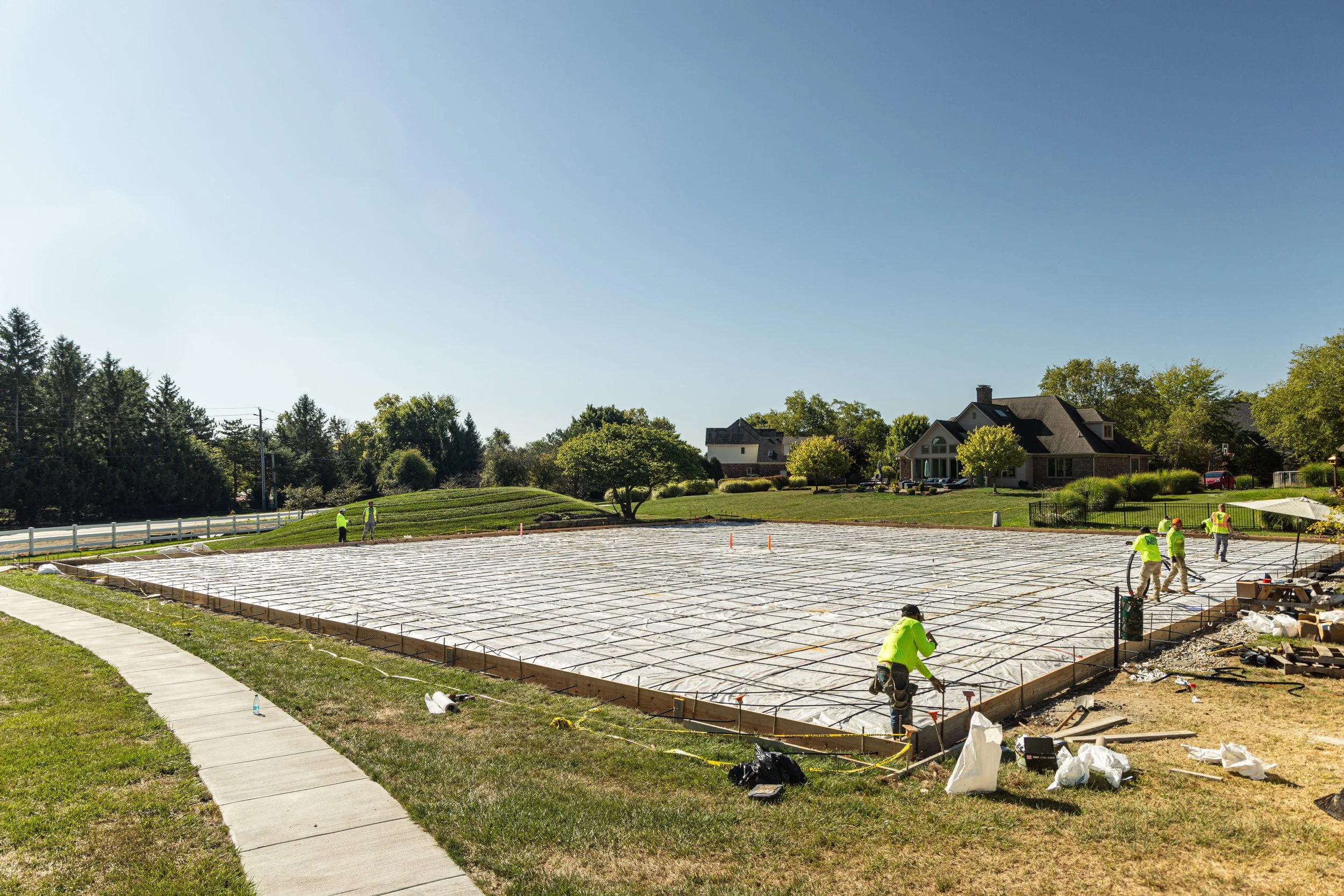 Construction workers laying foundation on a plot of land in a suburban neighborhood with finished houses and trees in the background.