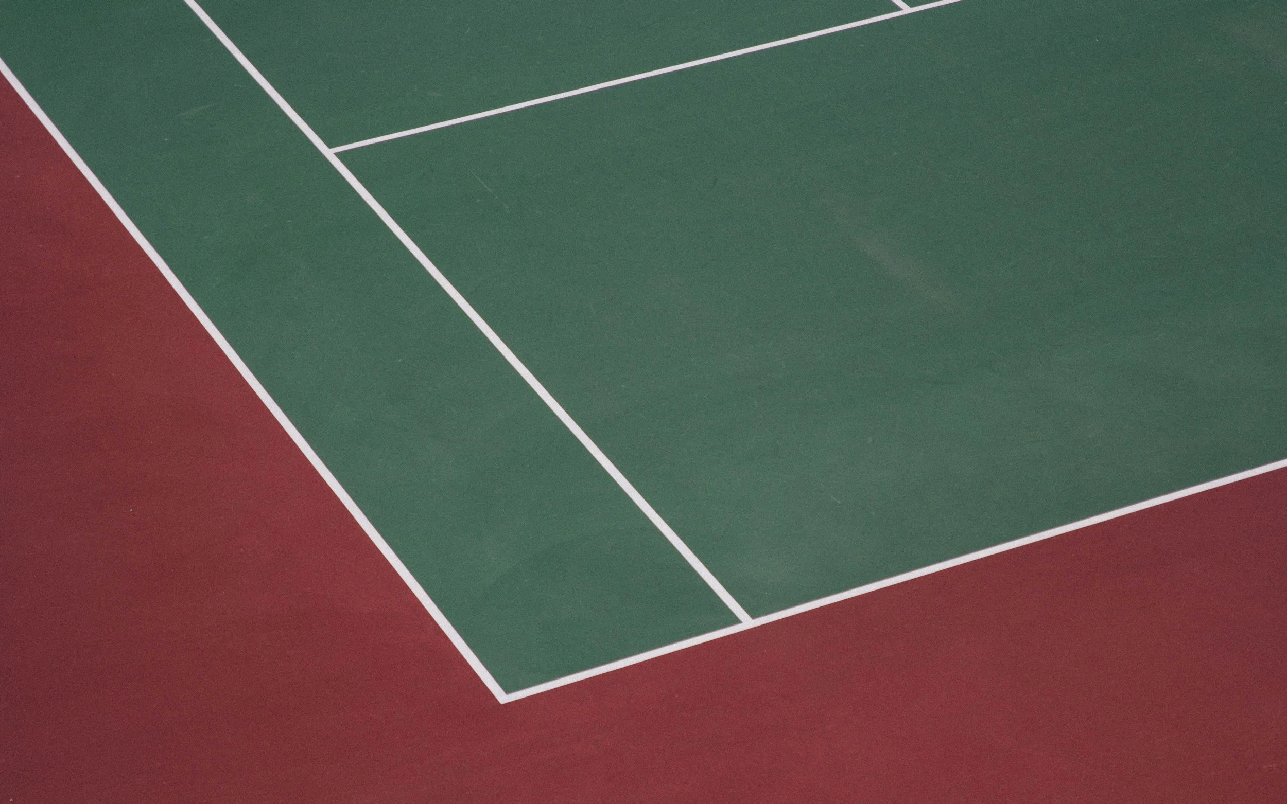 Close-up of a tennis court corner with green playing surface and red border, white boundary lines.
